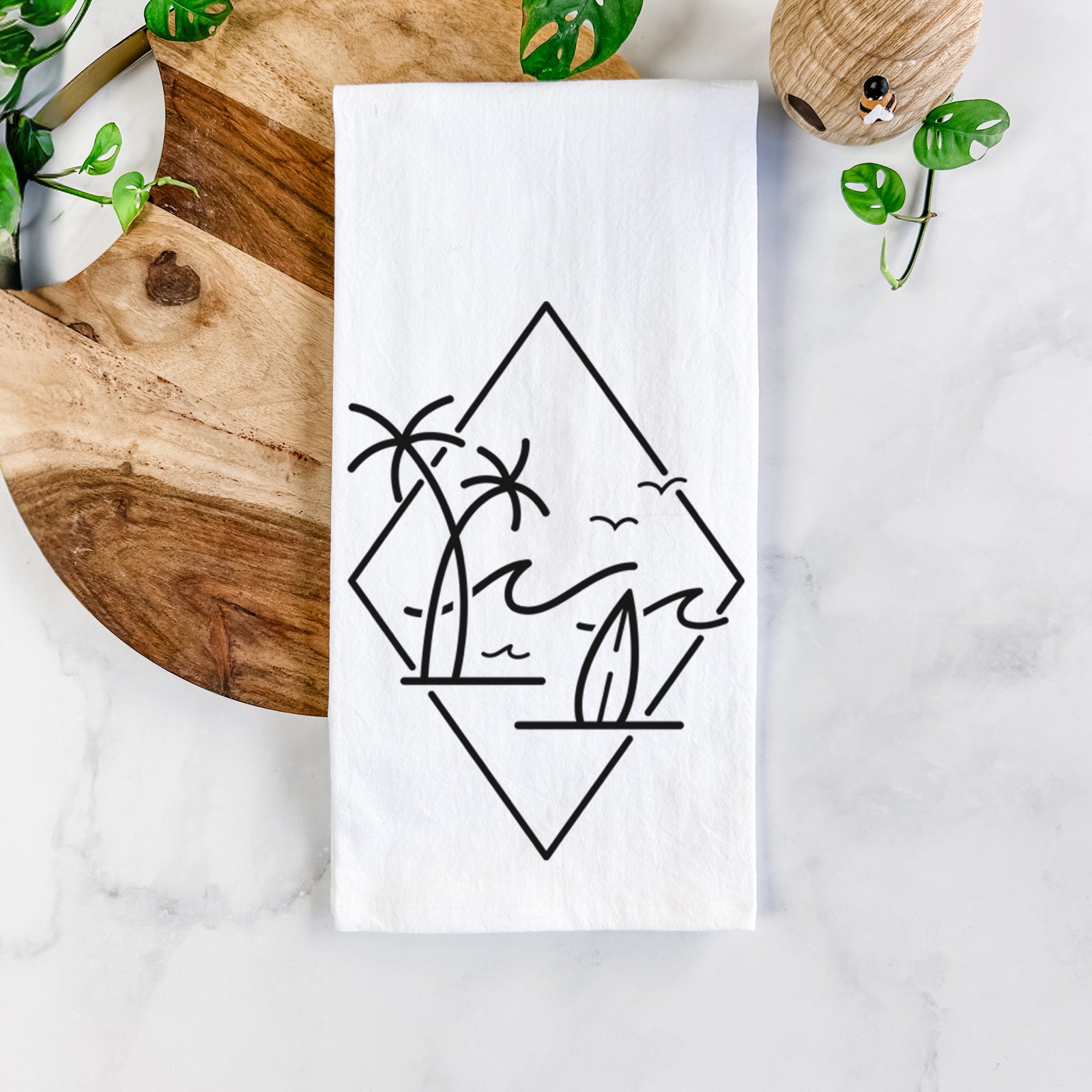 White cotton tea towel printed with black line art of palm trees, ocean waves, birds and a surfboard inside a diamond outline, on a marble counter beside a round wooden cutting board and greenery.