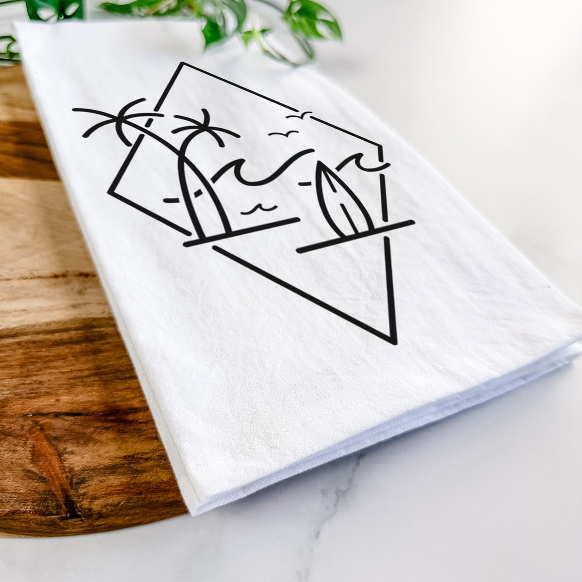 Close-up of a folded white tea towel on a wooden board, showing black line art of palm trees, ocean waves, birds and a surfboard inside a diamond outline.