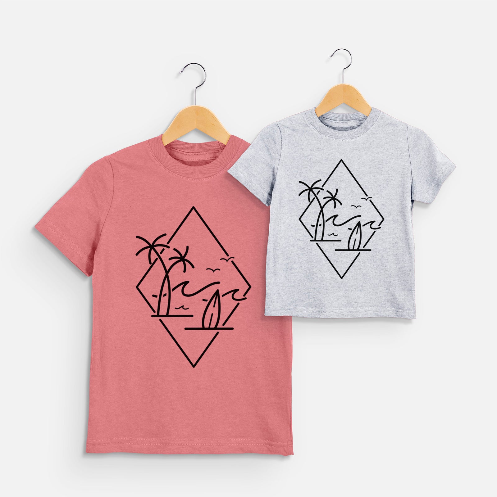 Two youth t-shirts in different colors on hangers, printed with black line art of palm trees, ocean waves, birds and a surfboard inside a diamond outline.