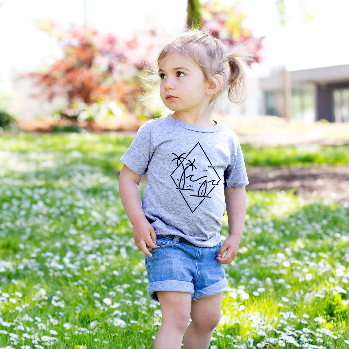 Toddler girl walking outdoors wearing a t-shirt printed with black line art of palm trees, ocean waves, birds and a surfboard inside a diamond outline.