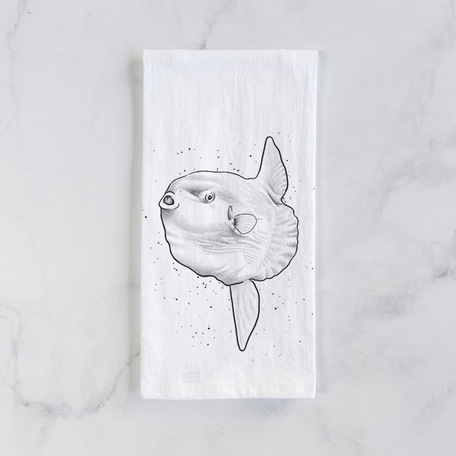 White cotton tea towel printed with a black ink illustration of an ocean sunfish swimming, on a marble counter beside a round wooden cutting board and greenery.