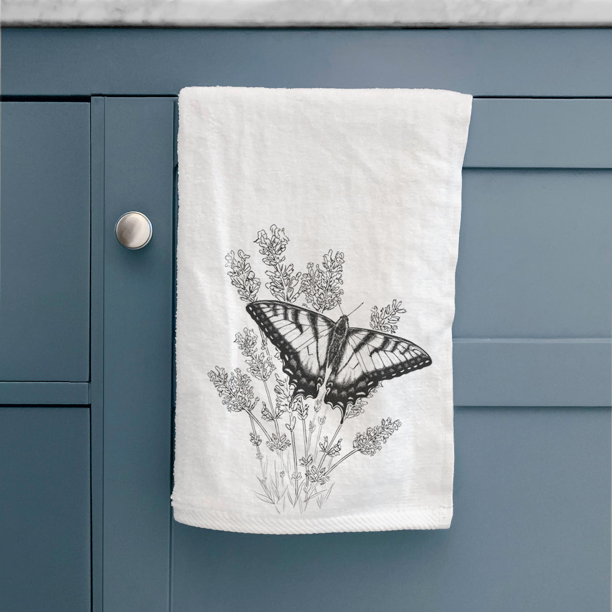 White hand towel printed with a black ink illustration of a swallowtail butterfly resting on lavender, hanging on a cabinet door.