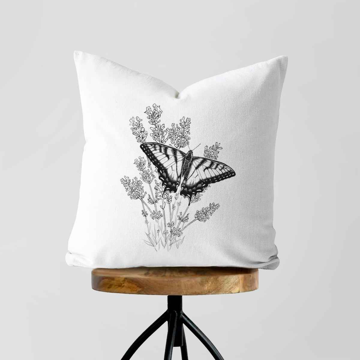 White pillow cover printed with a black ink illustration of a swallowtail butterfly resting on lavender, on a wooden stool.
