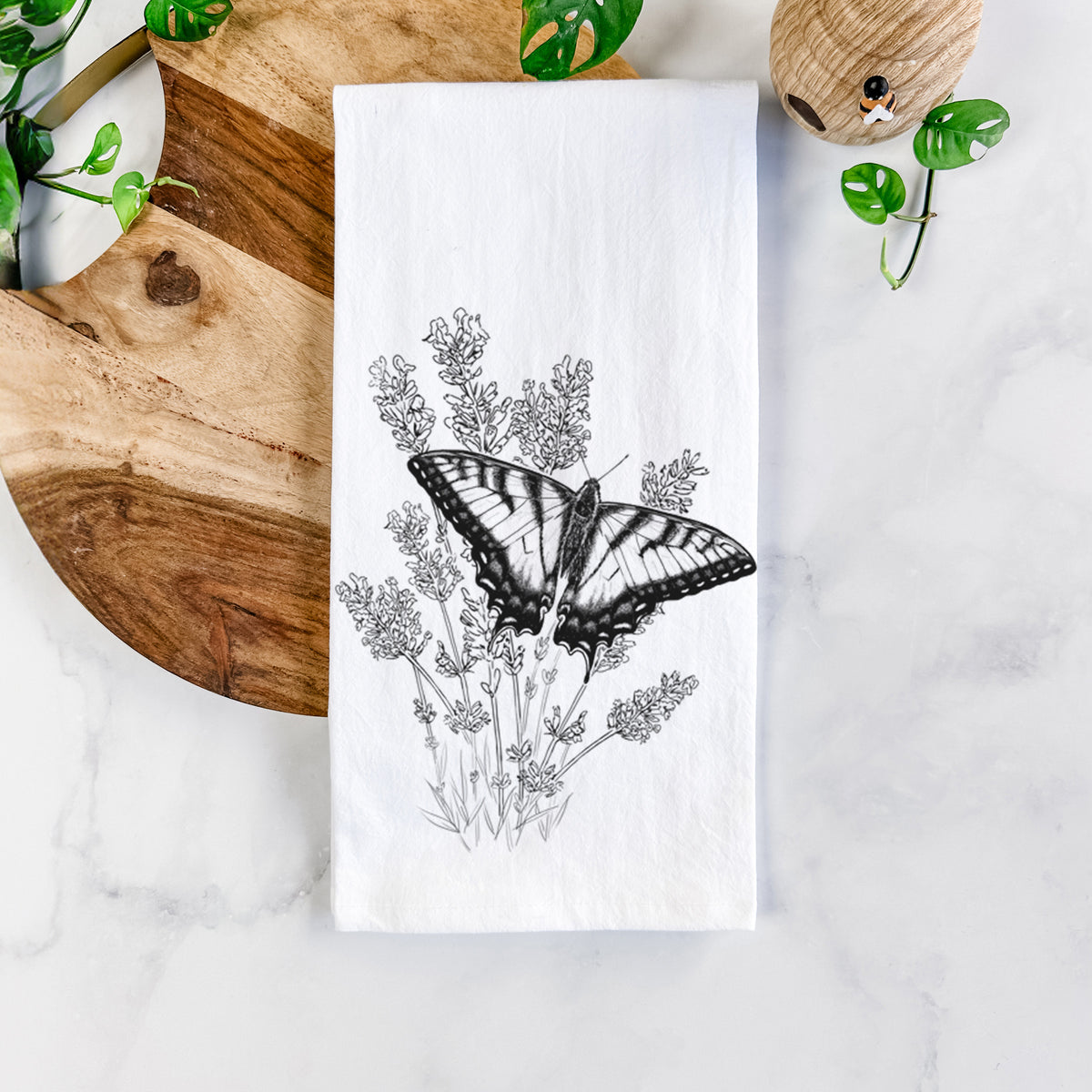 White cotton tea towel printed with a black ink illustration of a swallowtail butterfly resting on lavender, on a marble counter beside a round wooden cutting board and greenery.