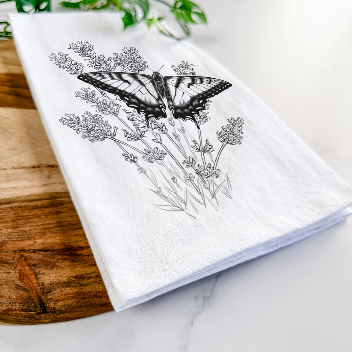 Close-up of a folded white tea towel on a wooden board, showing a black ink illustration of a swallowtail butterfly resting on lavender.