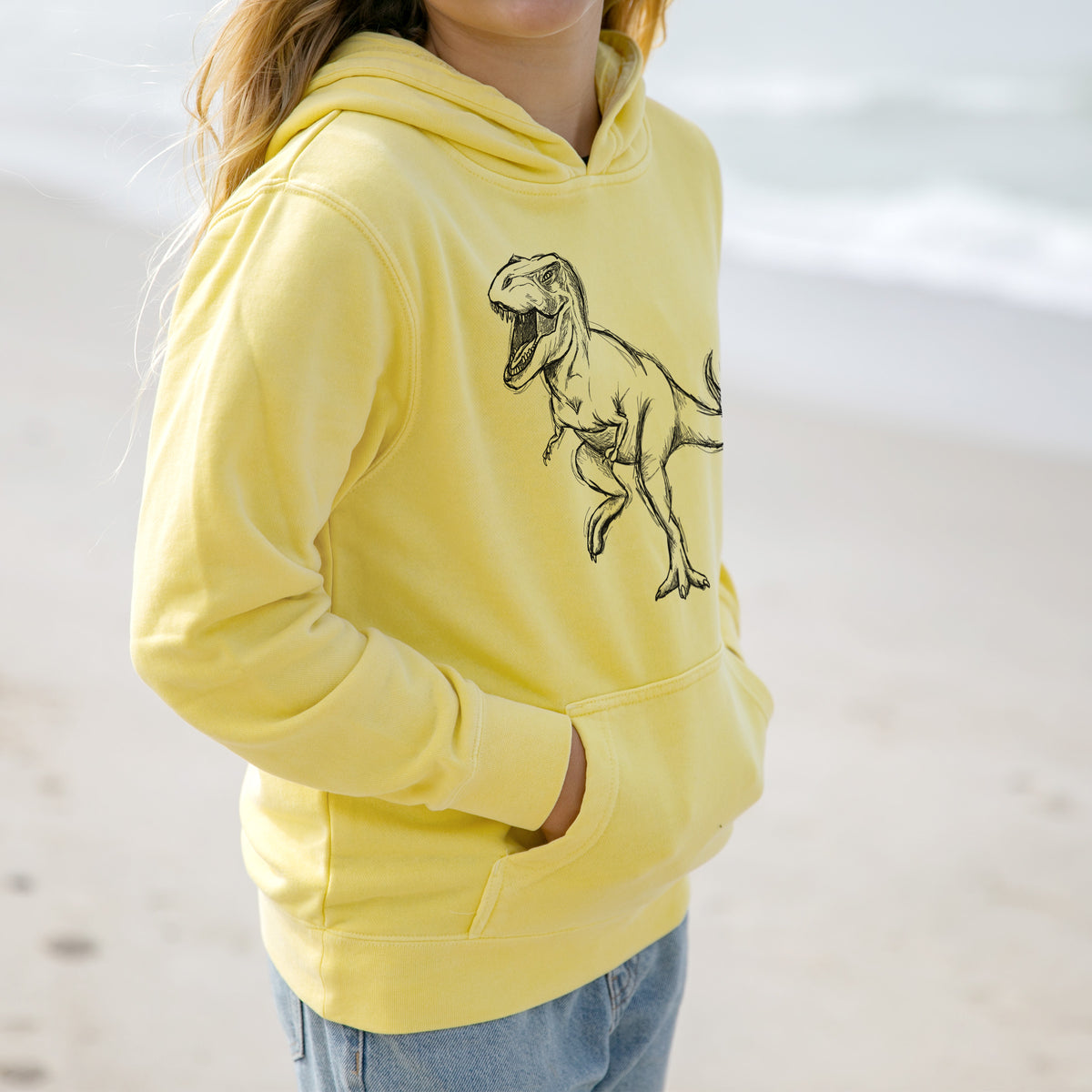 Girl wearing a youth pigment dyed hoodie printed with a black ink illustration of a Tyrannosaurus rex roaring.