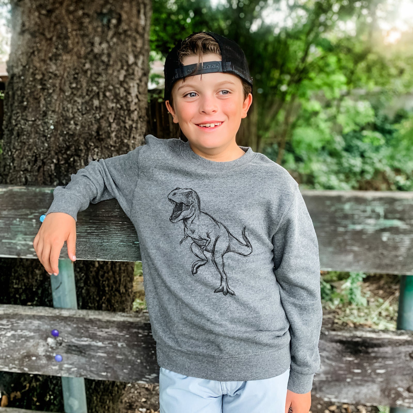 Boy wearing a youth crewneck sweatshirt printed with a black ink illustration of a Tyrannosaurus rex roaring, outdoors.