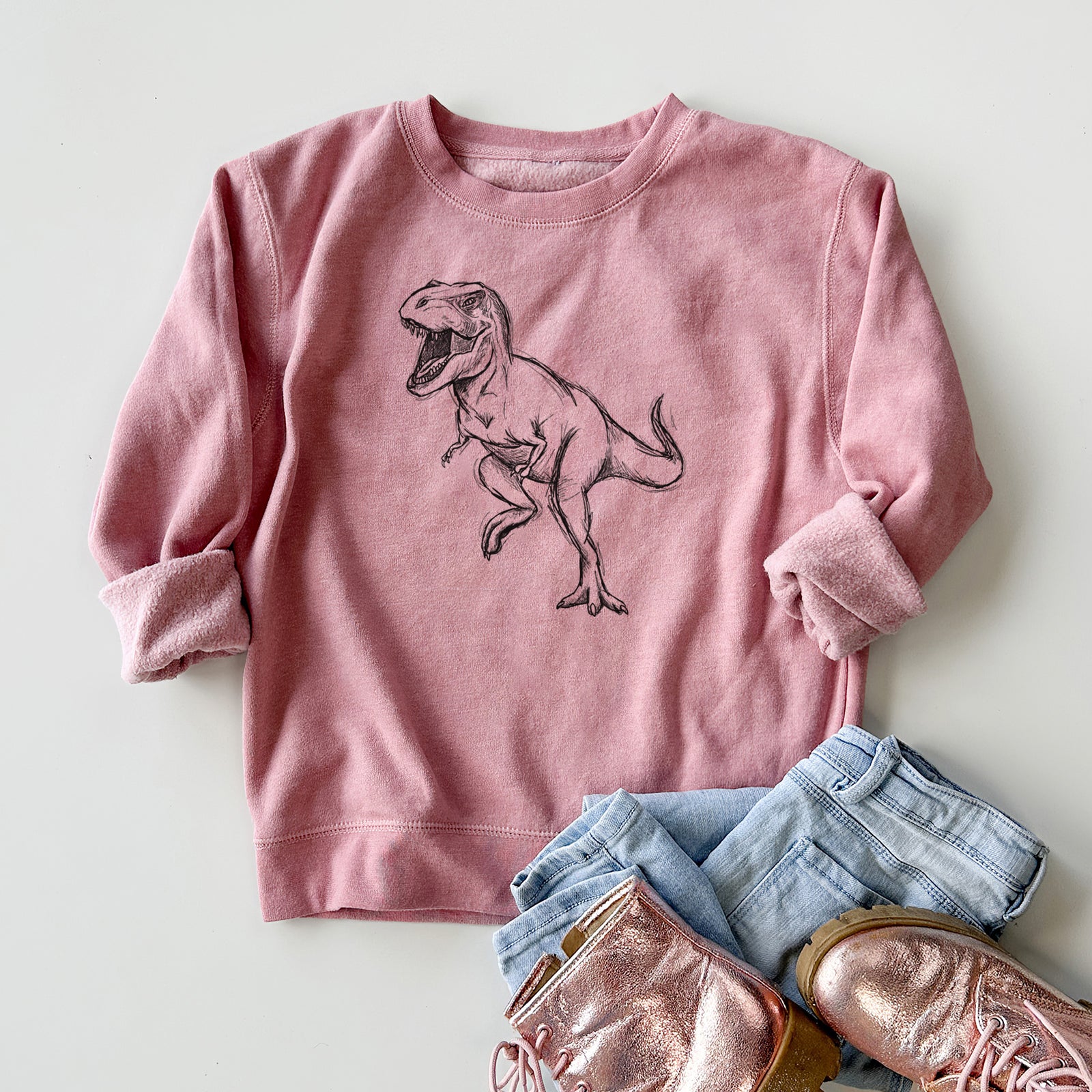 Boy wearing a youth crewneck sweatshirt printed with a black ink illustration of a Tyrannosaurus rex roaring, outdoors.