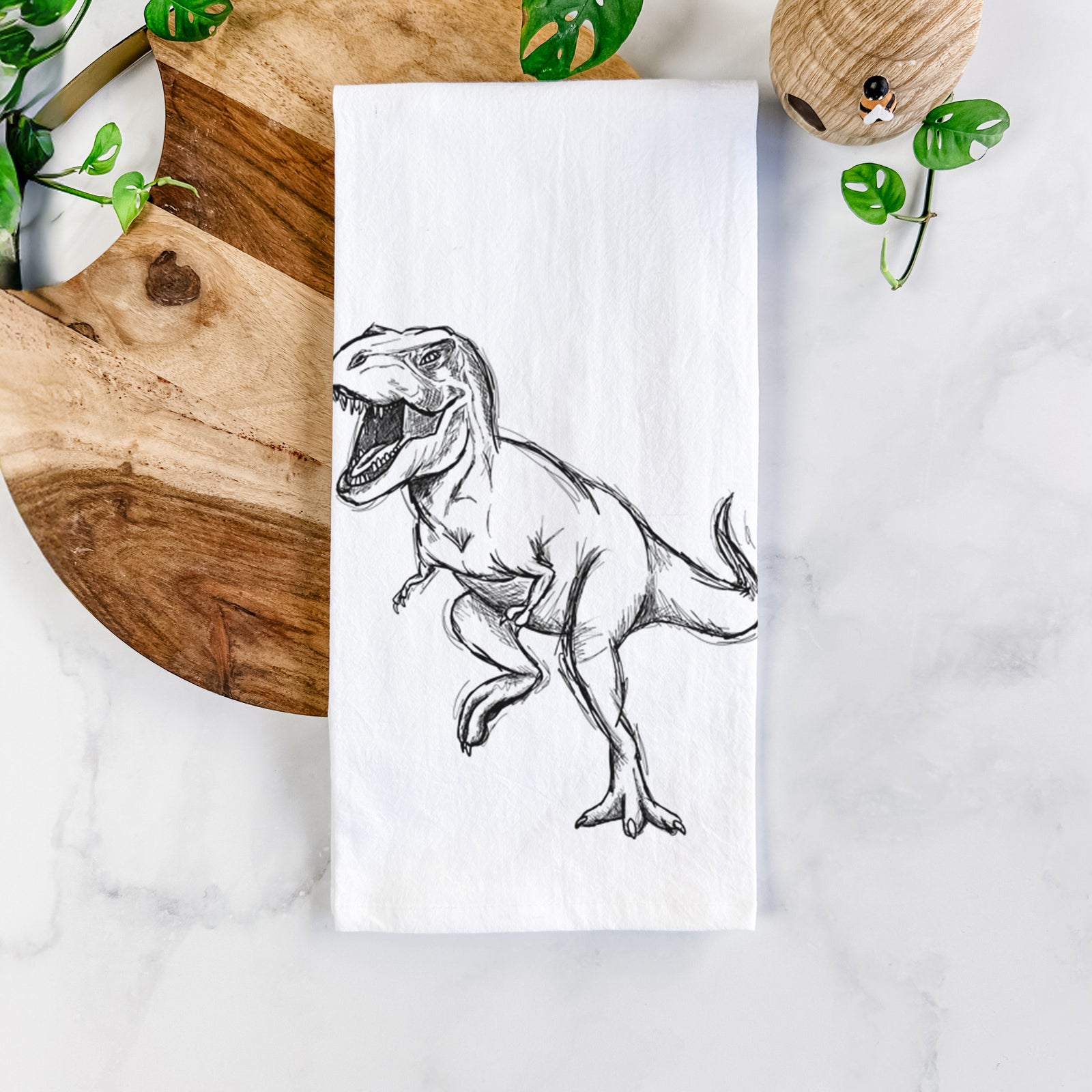 White cotton tea towel printed with a black ink illustration of a Tyrannosaurus rex roaring, on a marble counter beside a round wooden cutting board and greenery.