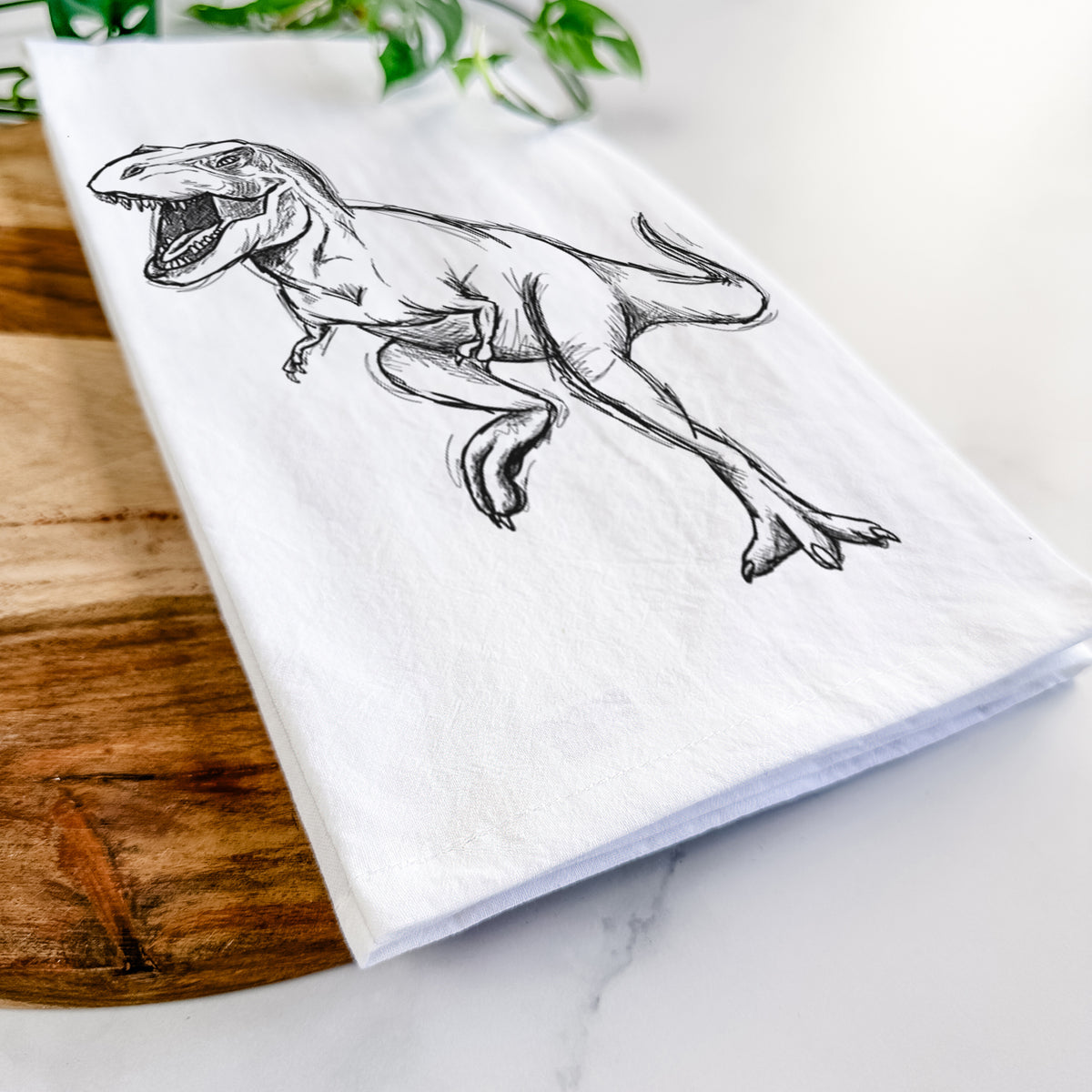 Close-up of a folded white tea towel on a wooden board, showing a black ink illustration of a Tyrannosaurus rex roaring.