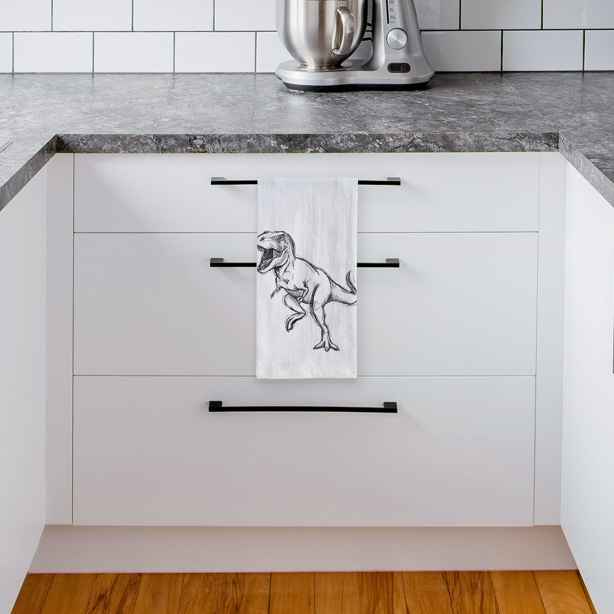 White tea towel printed with a black ink illustration of a Tyrannosaurus rex roaring, hanging from a drawer handle in a bright white kitchen.