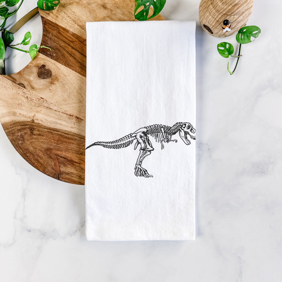 White cotton tea towel printed with a black ink illustration of a Tyrannosaurus rex skeleton, on a marble counter beside a round wooden cutting board and greenery.