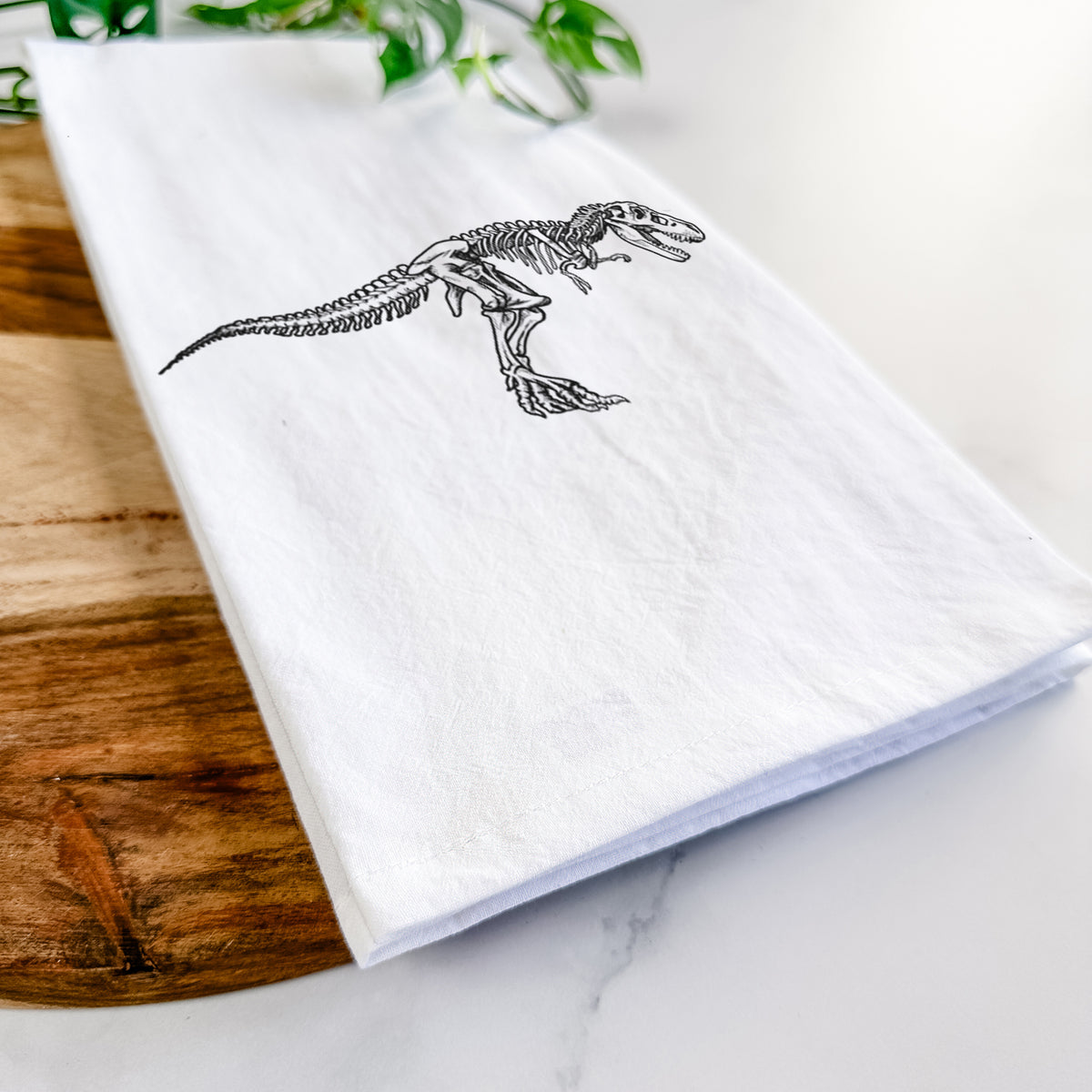 Close-up of a folded white tea towel on a wooden board, showing a black ink illustration of a Tyrannosaurus rex skeleton.