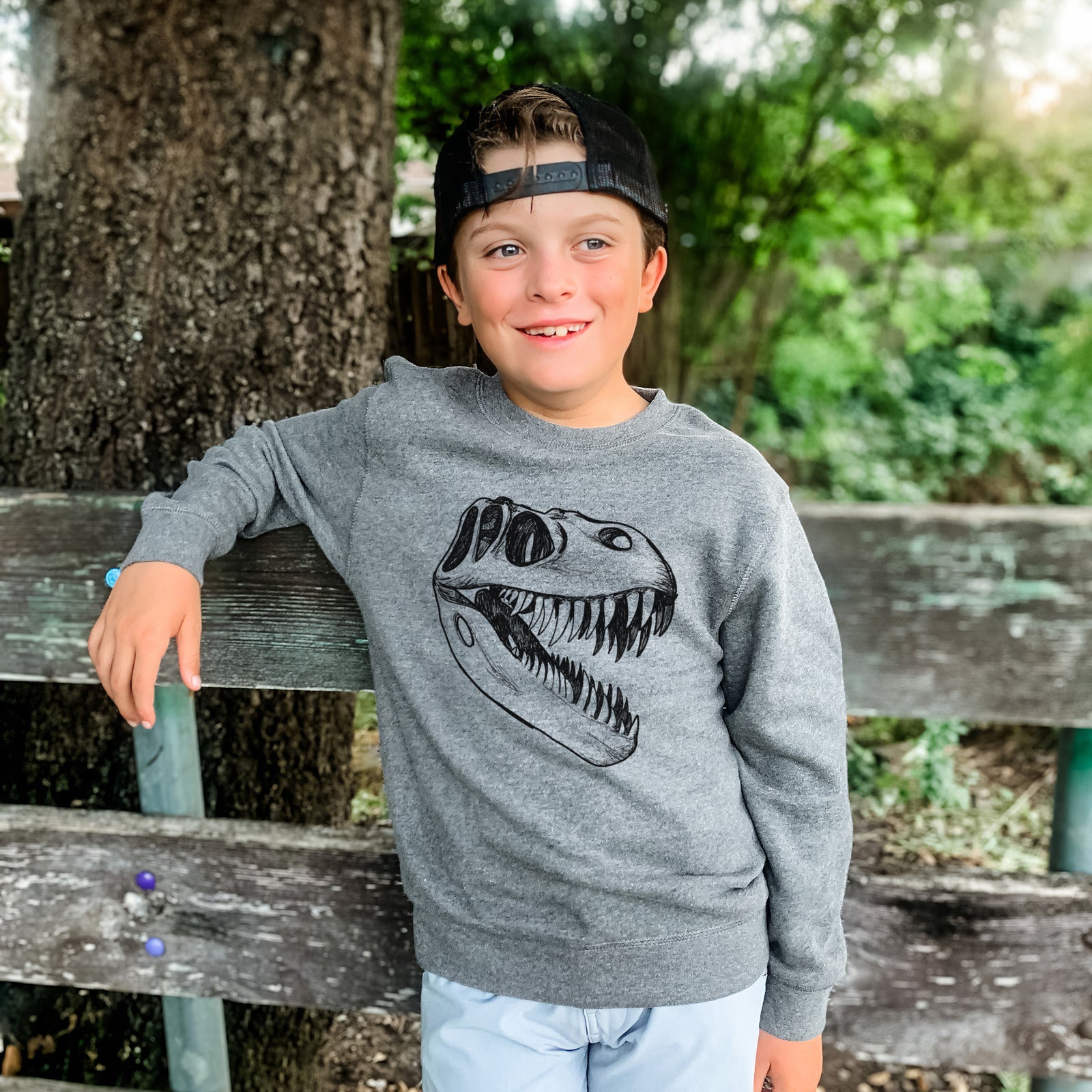 Boy wearing a youth crewneck sweatshirt printed with a black ink illustration of a Tyrannosaurus rex skull with sharp teeth, outdoors.