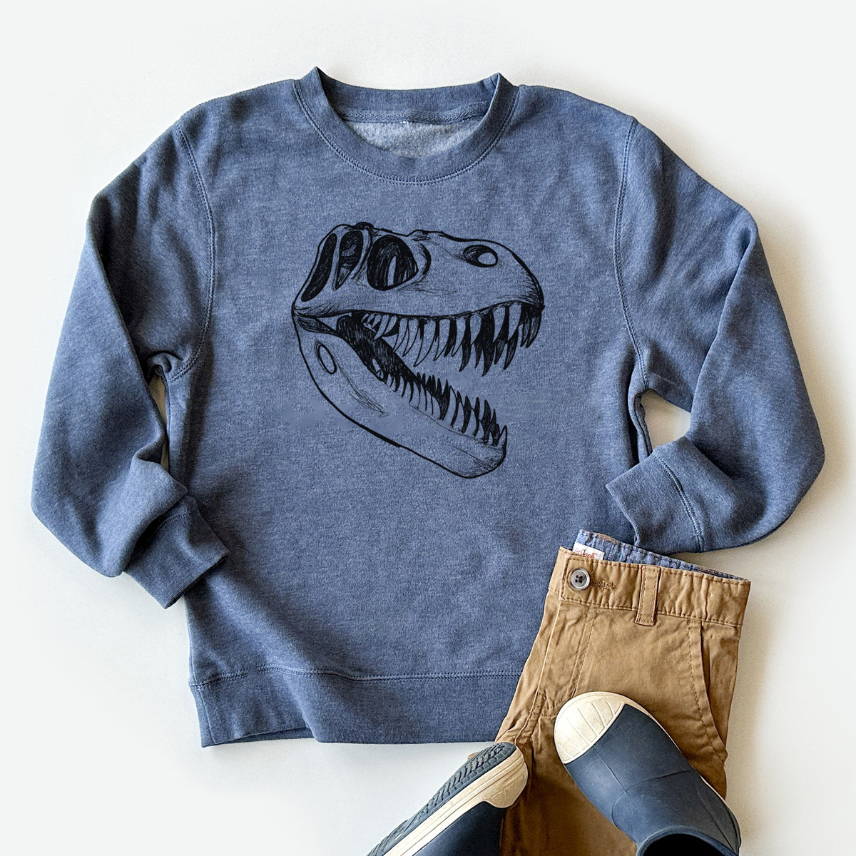 Youth crewneck sweatshirt printed with a black ink illustration of a Tyrannosaurus rex skull with sharp teeth, styled flat with khaki pants and shoes.