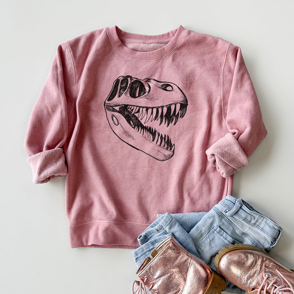 Youth crewneck sweatshirt printed with a black ink illustration of a Tyrannosaurus rex skull with sharp teeth, styled flat with jeans and boots.