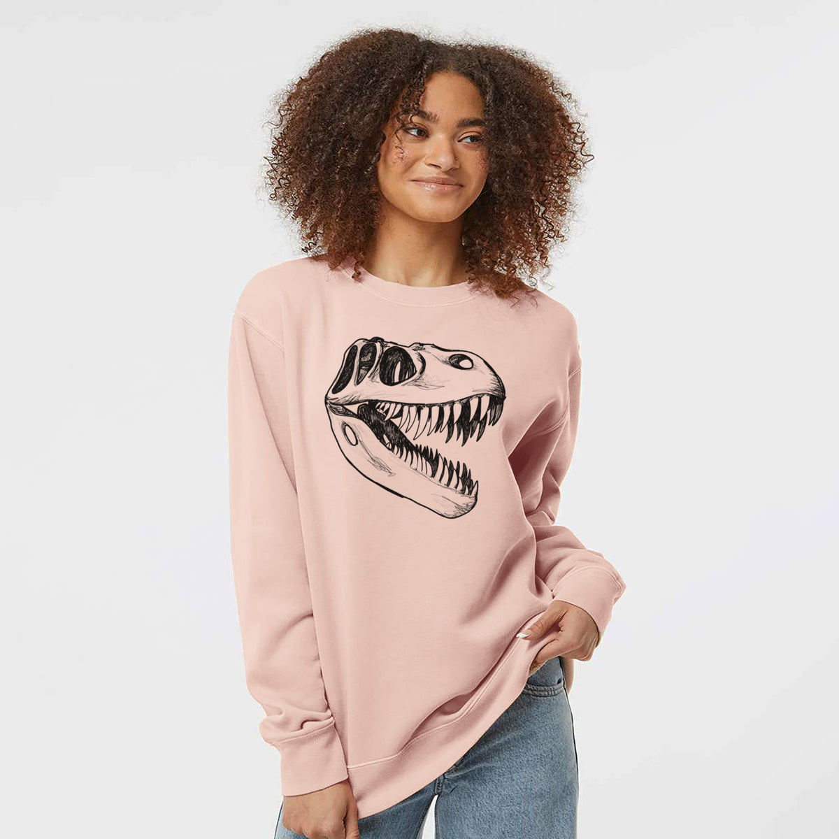 Model wearing a dusty pink pigment dyed crewneck sweatshirt printed with a black ink illustration of a Tyrannosaurus rex skull with sharp teeth.