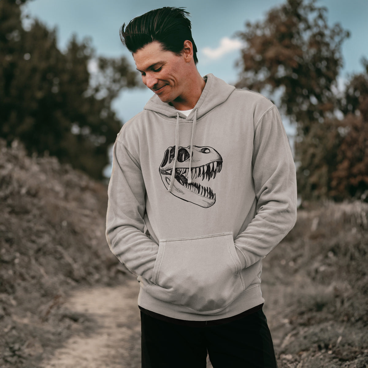 Man wearing a cement grey pigment dyed hoodie printed with a black ink illustration of a Tyrannosaurus rex skull with sharp teeth.