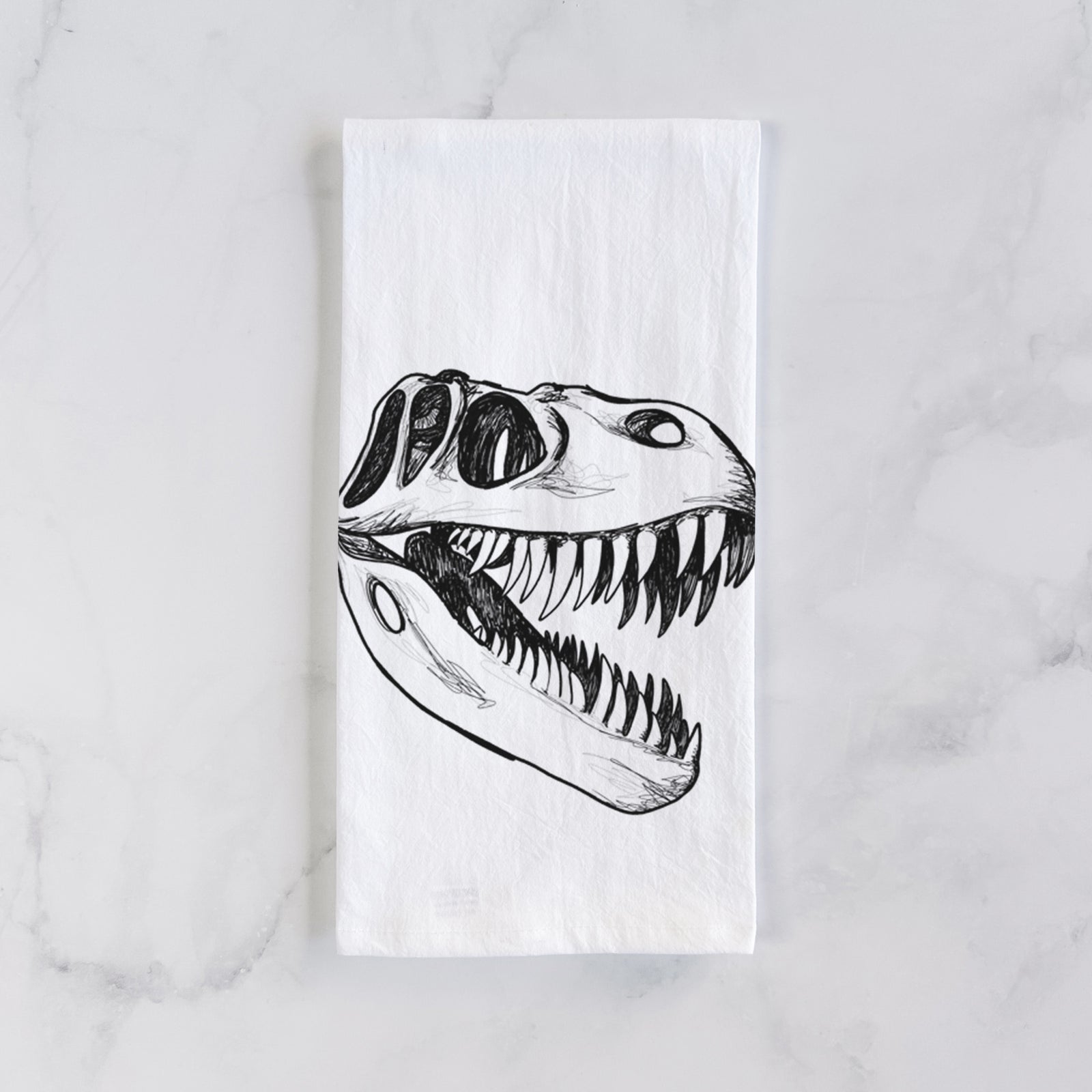 White cotton tea towel printed with a black ink illustration of a Tyrannosaurus rex skull with sharp teeth, on a marble counter beside a round wooden cutting board and greenery.