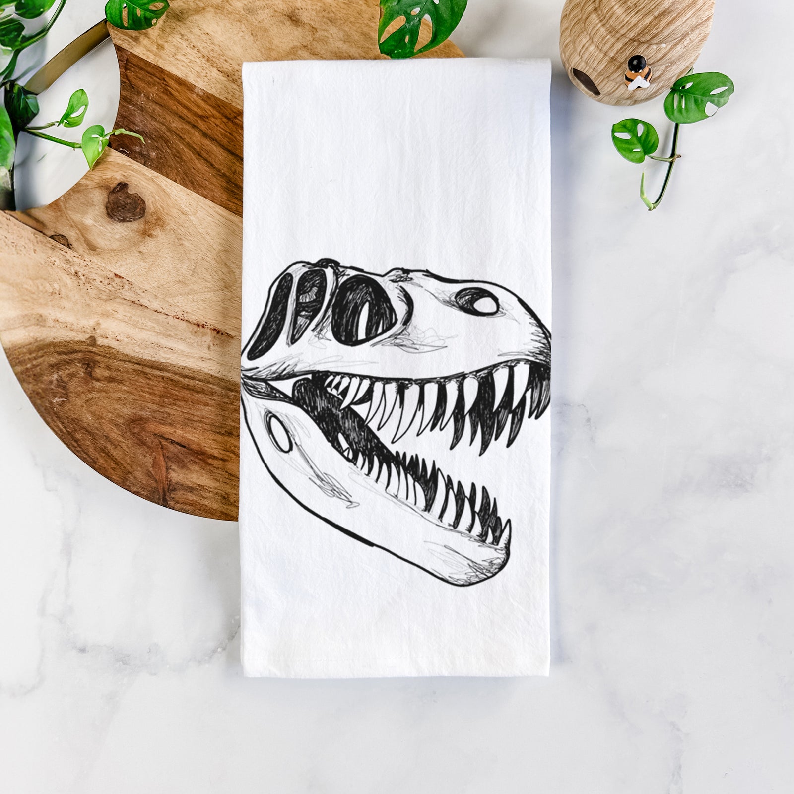 White cotton tea towel printed with a black ink illustration of a Tyrannosaurus rex skull with sharp teeth, on a marble counter beside a round wooden cutting board and greenery.