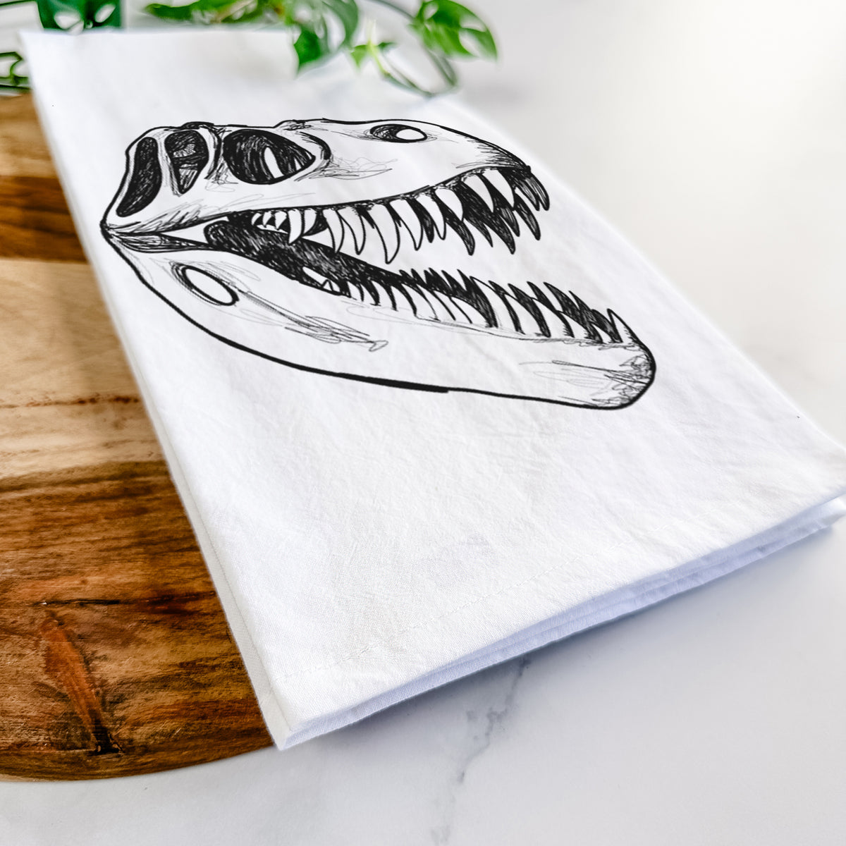 Close-up of a folded white tea towel on a wooden board, showing a black ink illustration of a Tyrannosaurus rex skull with sharp teeth.