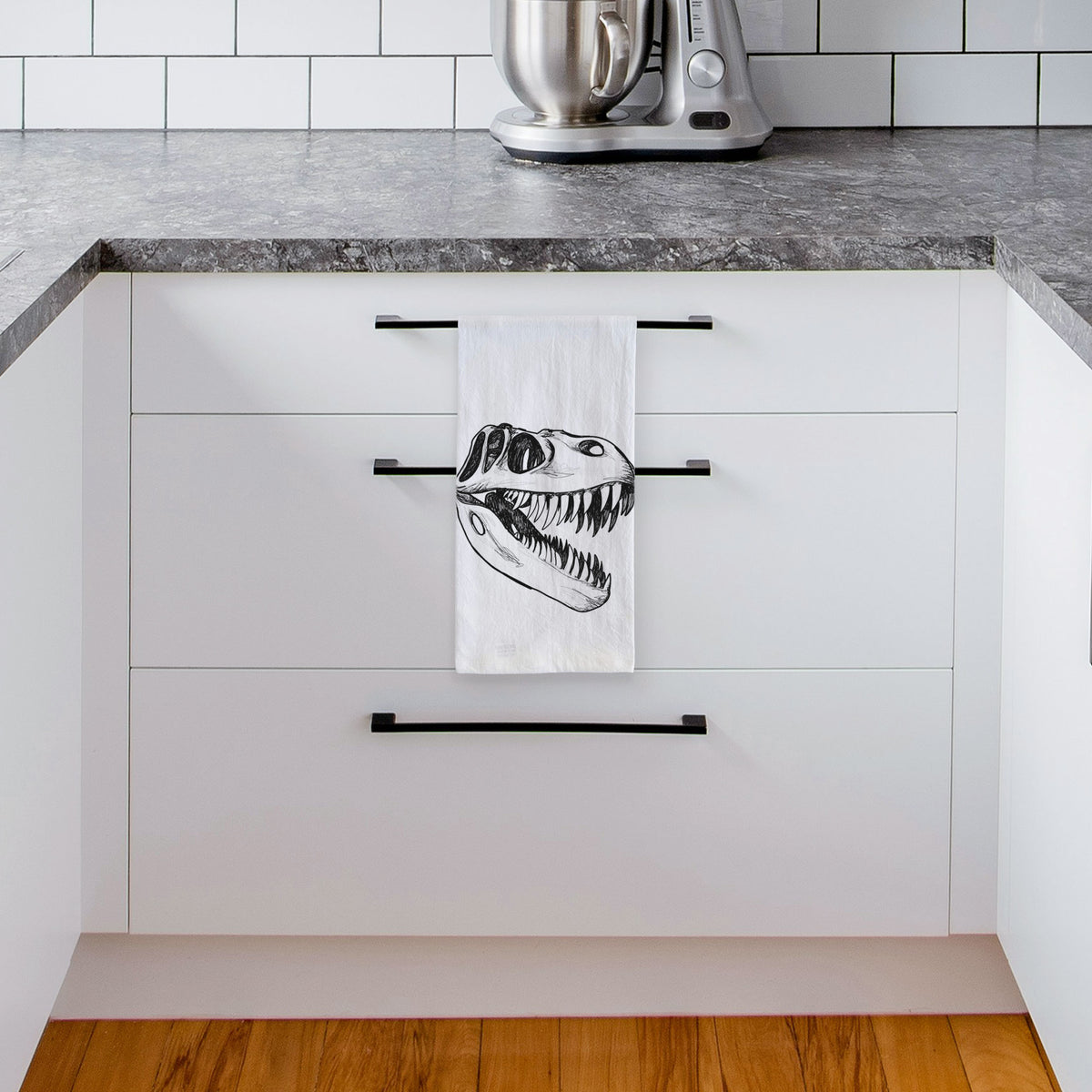 White tea towel printed with a black ink illustration of a Tyrannosaurus rex skull with sharp teeth, hanging from a drawer handle in a bright white kitchen.