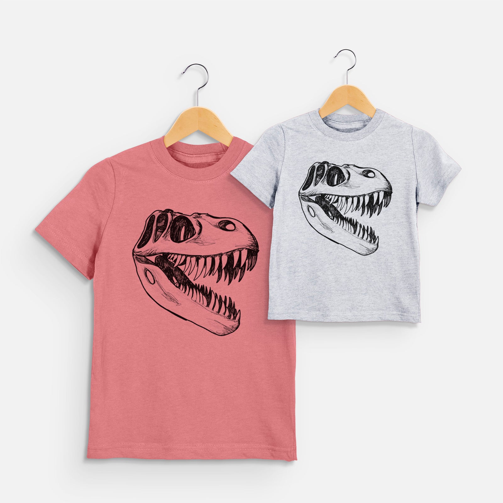 Two youth t-shirts in different colors on hangers, printed with a black ink illustration of a Tyrannosaurus rex skull with sharp teeth.