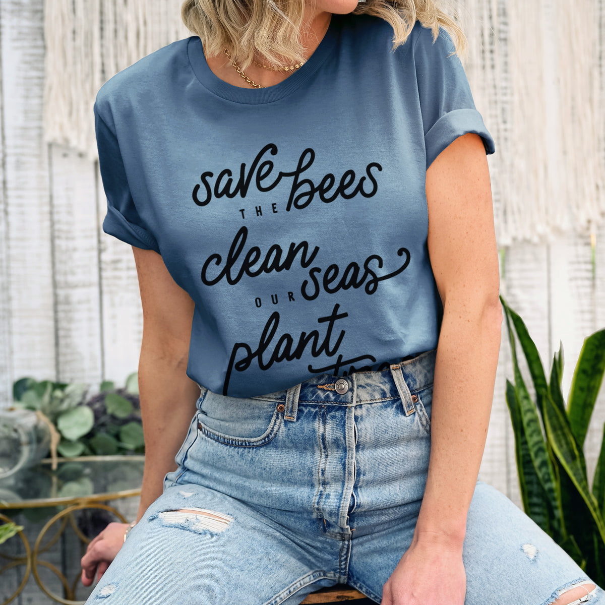 Woman wearing a steel blue lightweight cotton t-shirt printed with black hand-lettered words reading Save the Bees, Clean Our Seas, Plant More Trees.