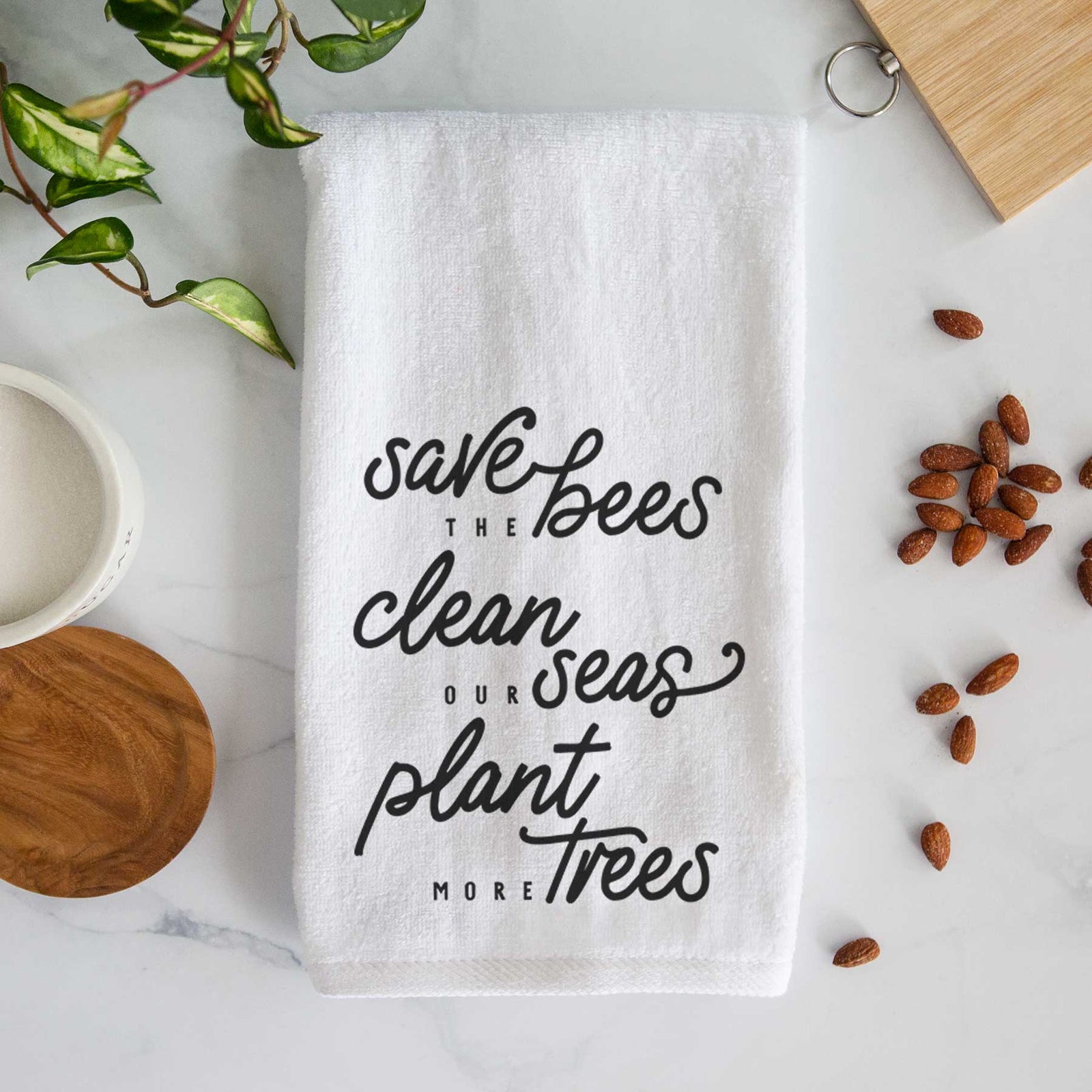 White hand towel printed with black hand-lettered words reading Save the Bees, Clean Our Seas, Plant More Trees.