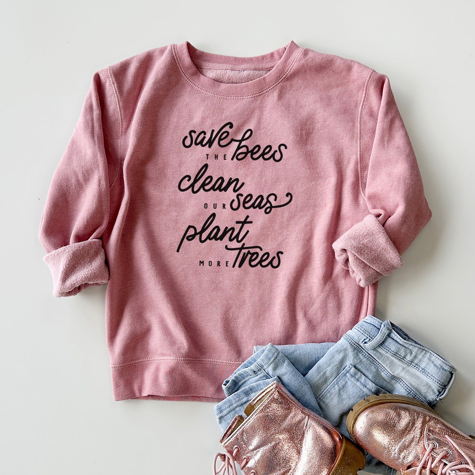Boy wearing a youth crewneck sweatshirt printed with black hand-lettered words reading Save the Bees, Clean Our Seas, Plant More Trees, outdoors.