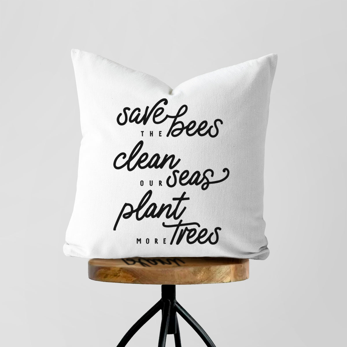 White pillow cover printed with black hand-lettered words reading Save the Bees, Clean Our Seas, Plant More Trees, on a wooden stool.
