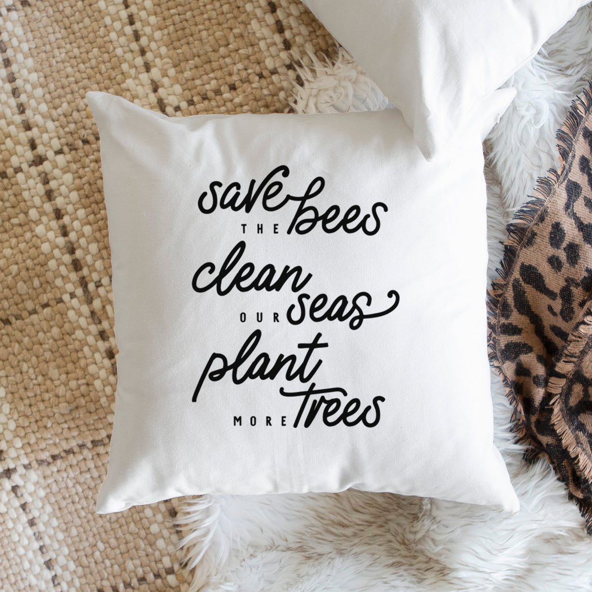 White pillow cover printed with black hand-lettered words reading Save the Bees, Clean Our Seas, Plant More Trees, on a woven chair.