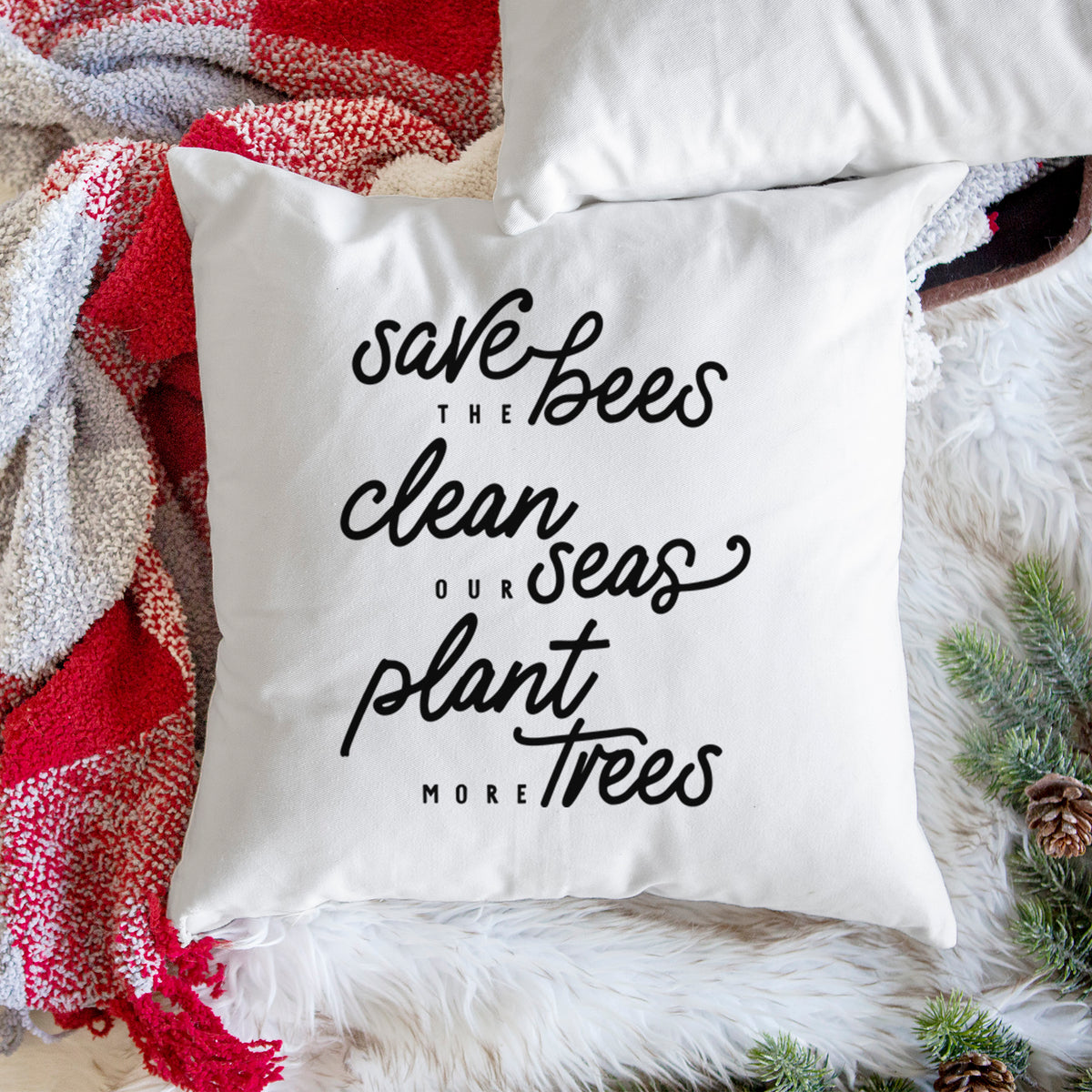 White pillow cover printed with black hand-lettered words reading Save the Bees, Clean Our Seas, Plant More Trees, on a woven chair.