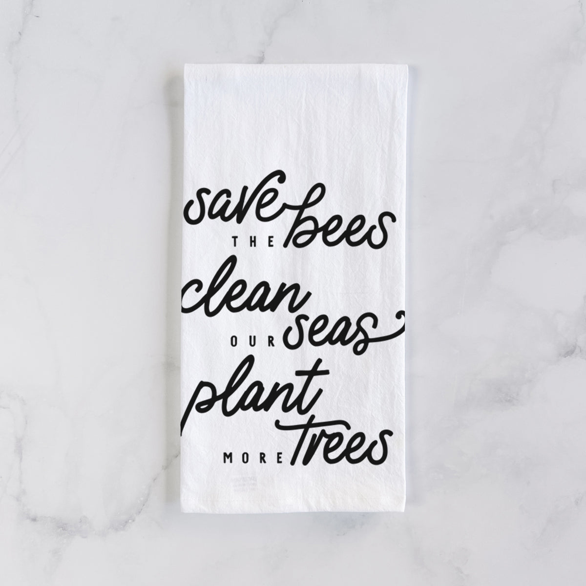 White cotton tea towel laid flat on marble, printed with black hand-lettered words reading Save the Bees, Clean Our Seas, Plant More Trees.
