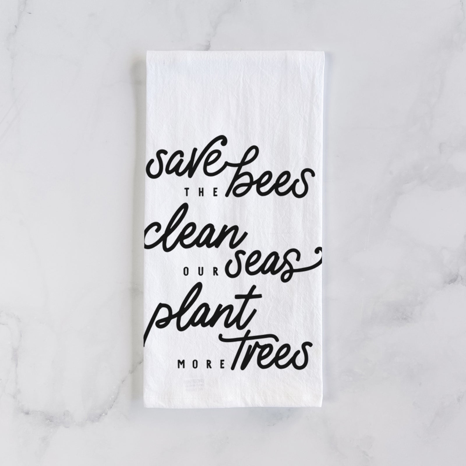 White cotton tea towel printed with black hand-lettered words reading Save the Bees, Clean Our Seas, Plant More Trees, on a marble counter beside a round wooden cutting board and greenery.