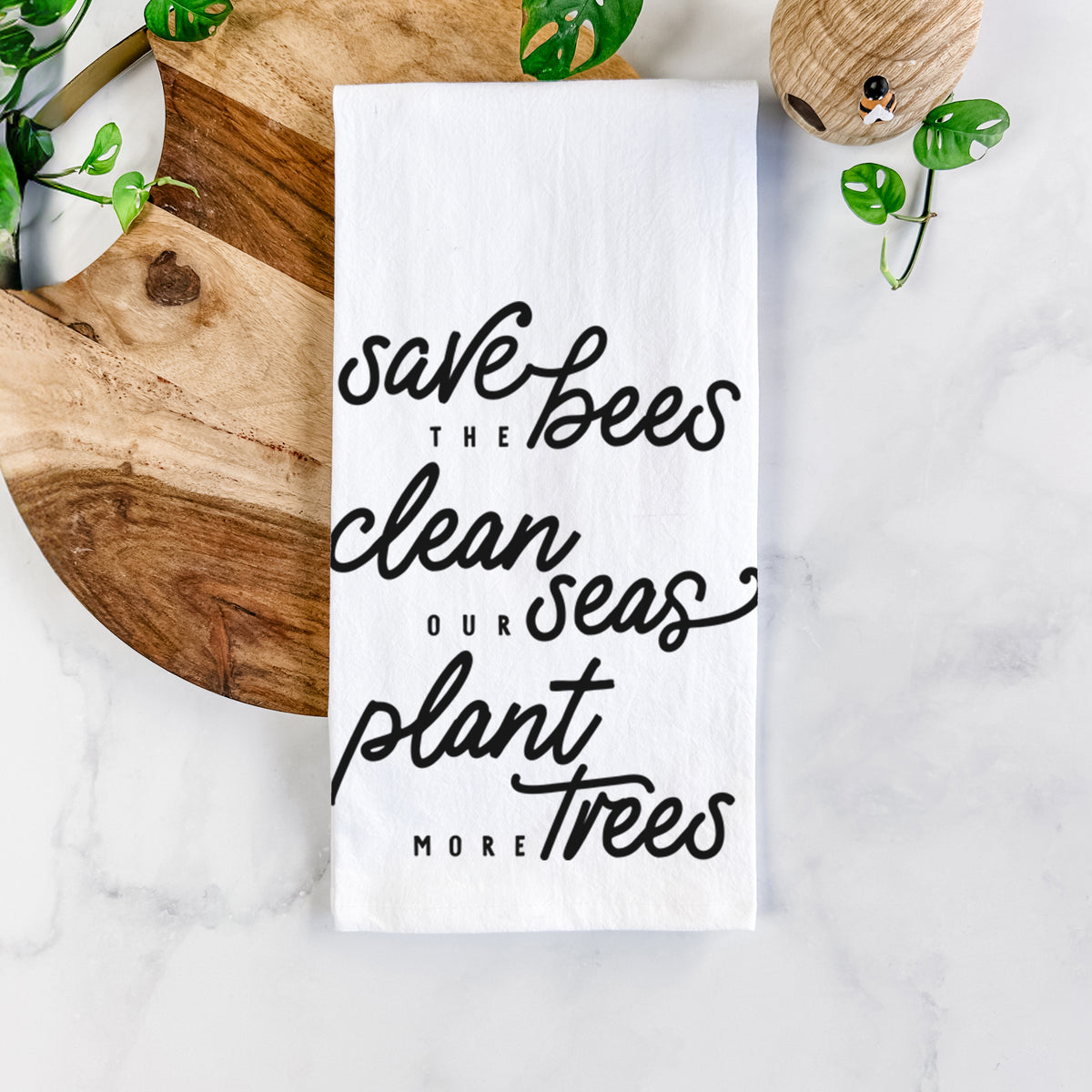 White cotton tea towel printed with black hand-lettered words reading Save the Bees, Clean Our Seas, Plant More Trees, on a marble counter beside a round wooden cutting board and greenery.