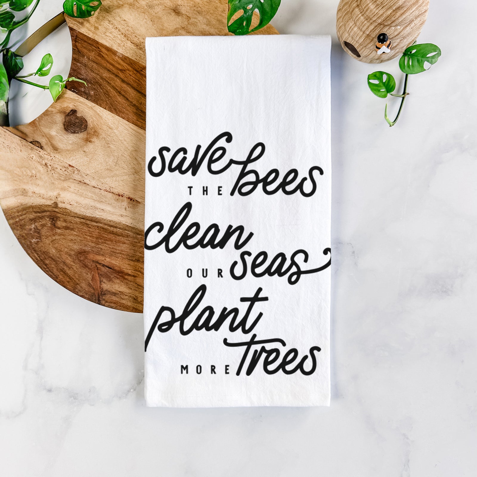 White cotton tea towel printed with black hand-lettered words reading Save the Bees, Clean Our Seas, Plant More Trees, on a marble counter beside a round wooden cutting board and greenery.