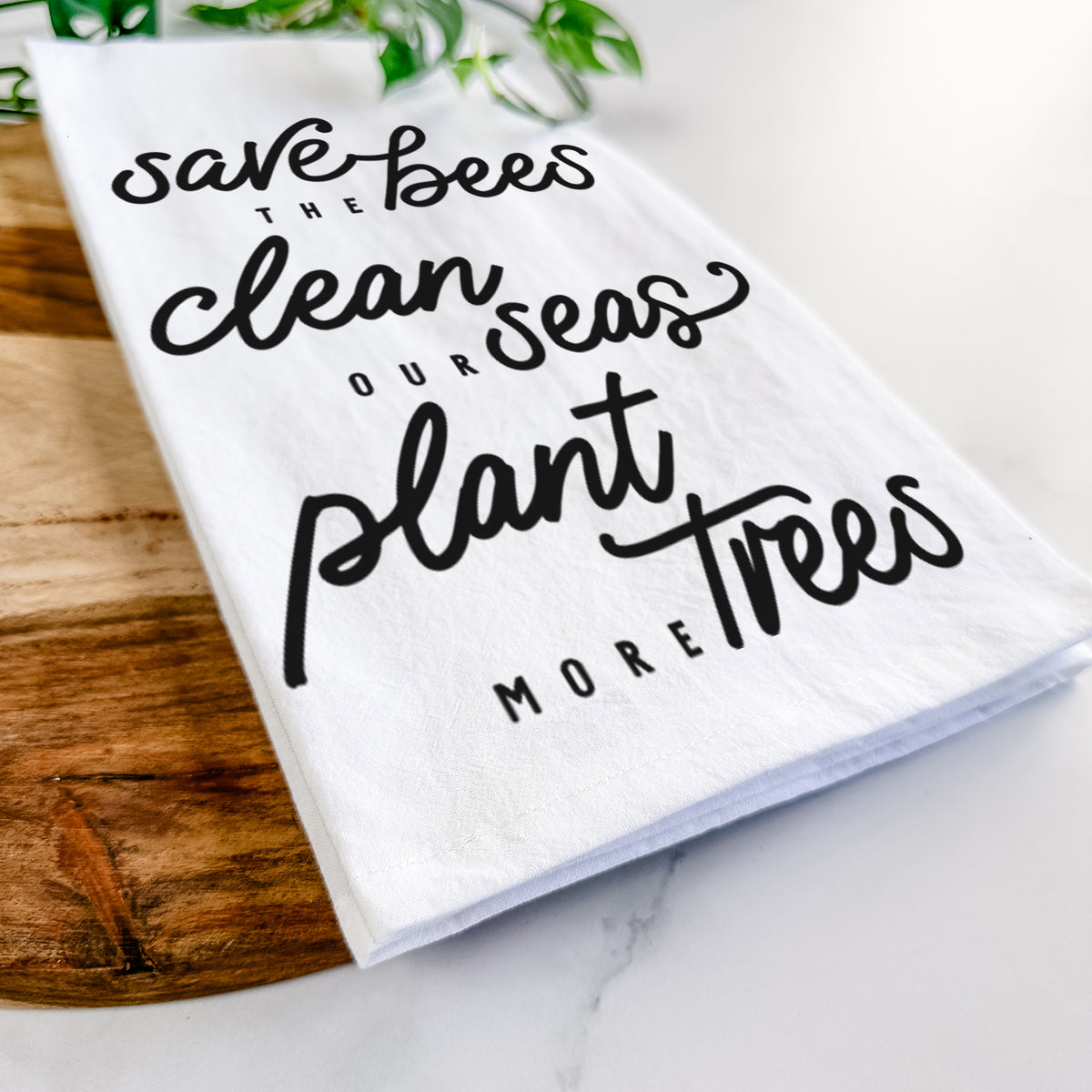 Close-up of a folded white tea towel on a wooden board, showing black hand-lettered words reading Save the Bees, Clean Our Seas, Plant More Trees.