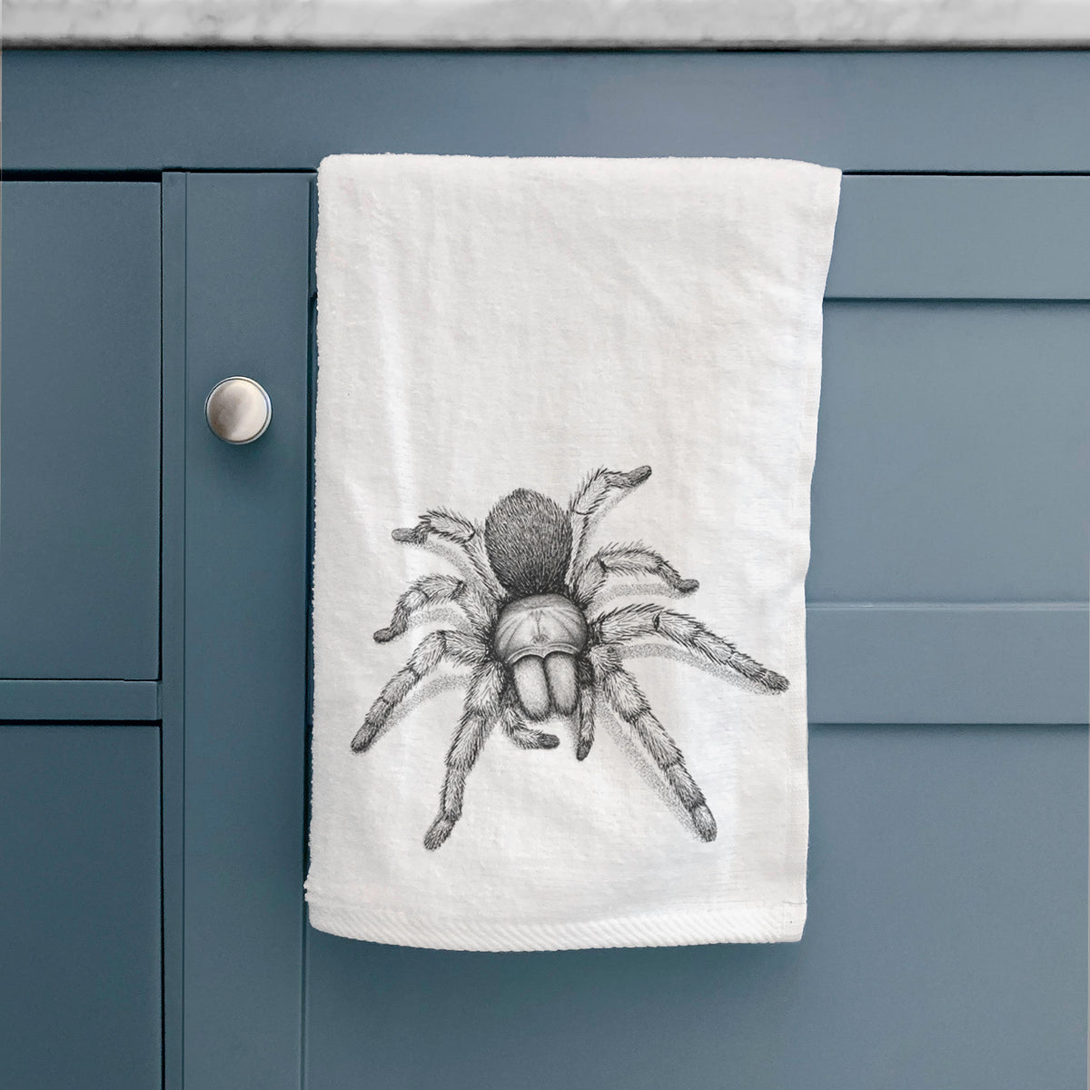 White hand towel printed with a detailed black ink illustration of a desert blonde tarantula, hanging on a cabinet door.