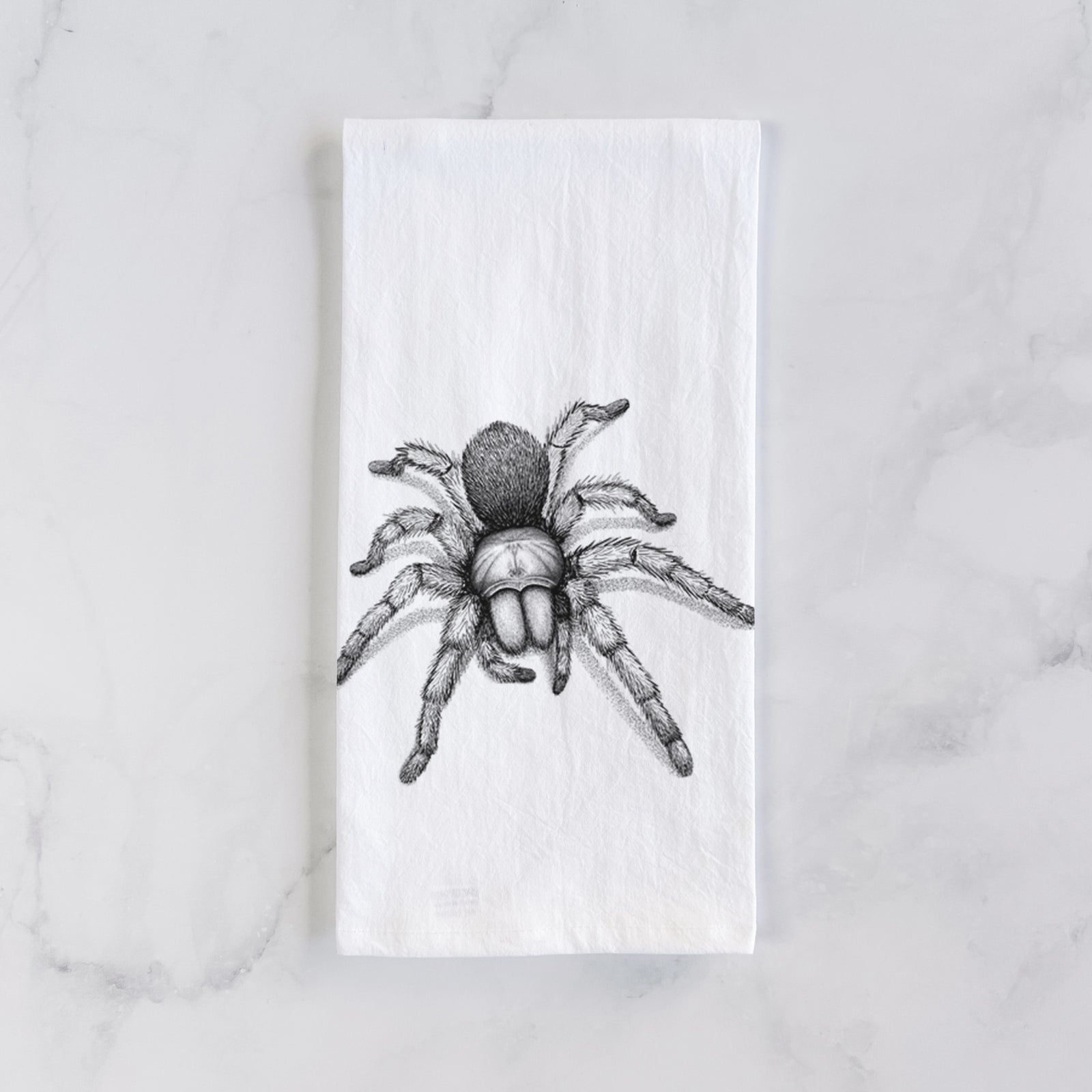 White cotton tea towel printed with a detailed black ink illustration of a desert blonde tarantula, on a marble counter beside a round wooden cutting board and greenery.