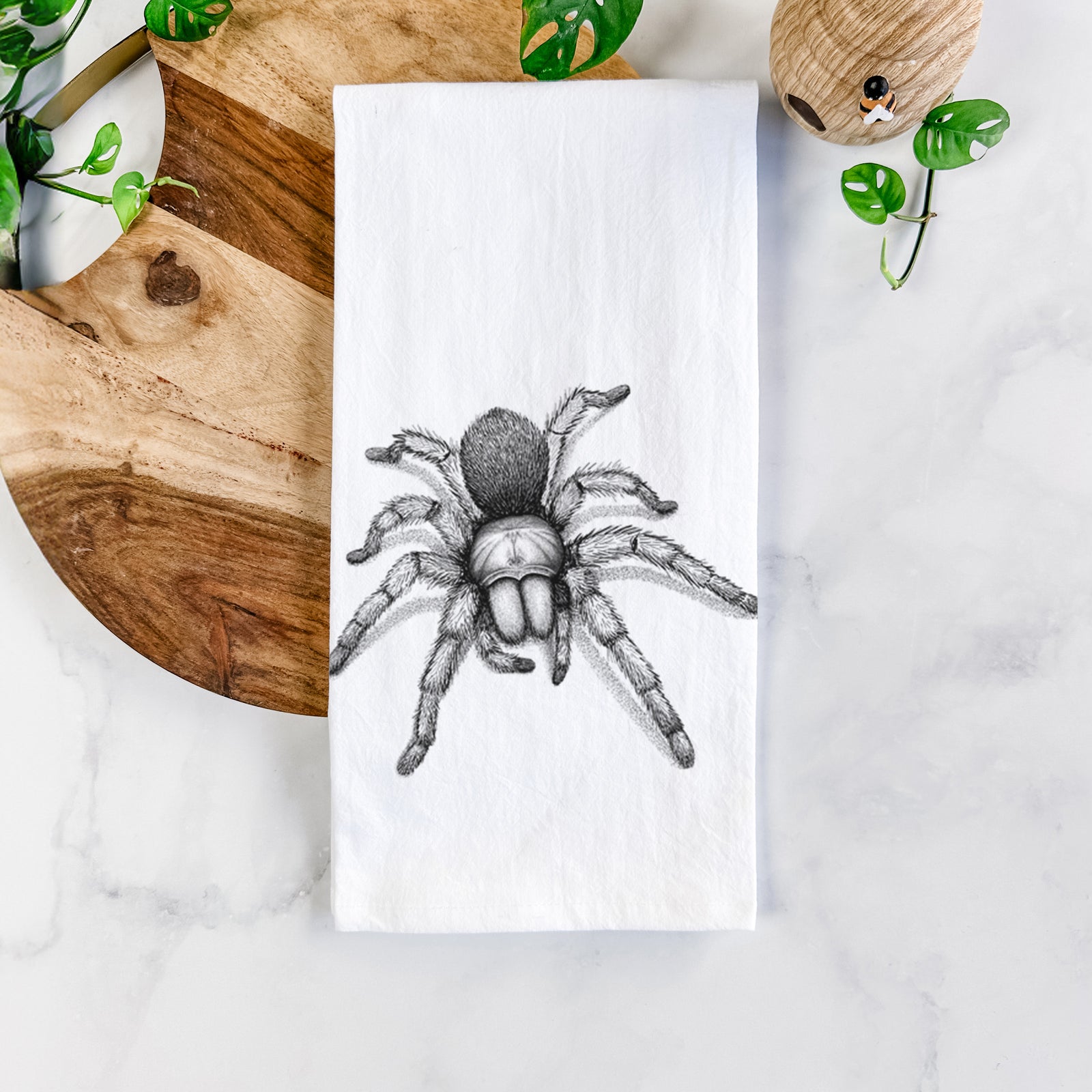White cotton tea towel printed with a detailed black ink illustration of a desert blonde tarantula, on a marble counter beside a round wooden cutting board and greenery.