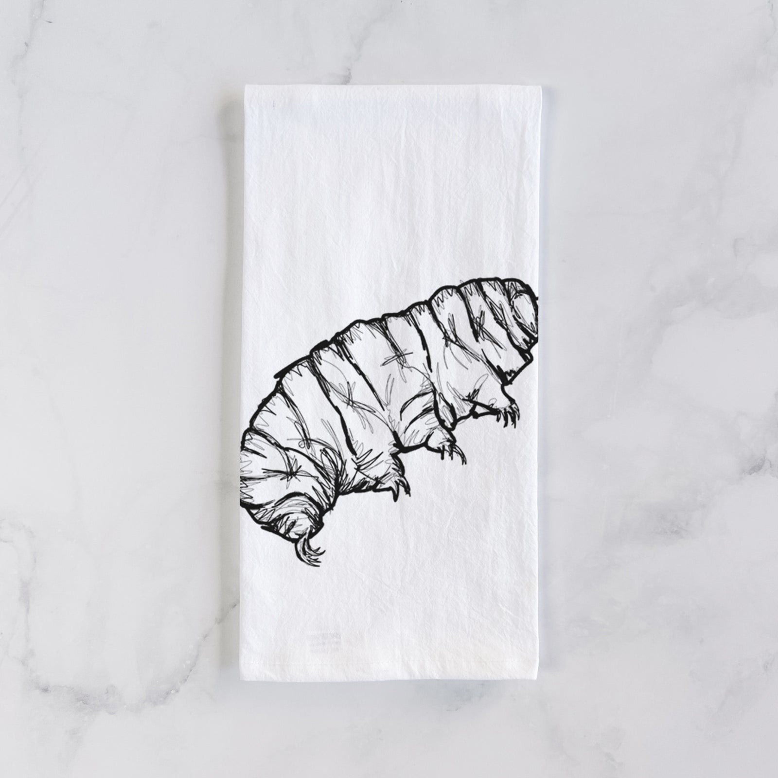White cotton tea towel printed with a detailed black ink illustration of a tardigrade, or water bear, on a marble counter beside a round wooden cutting board and greenery.