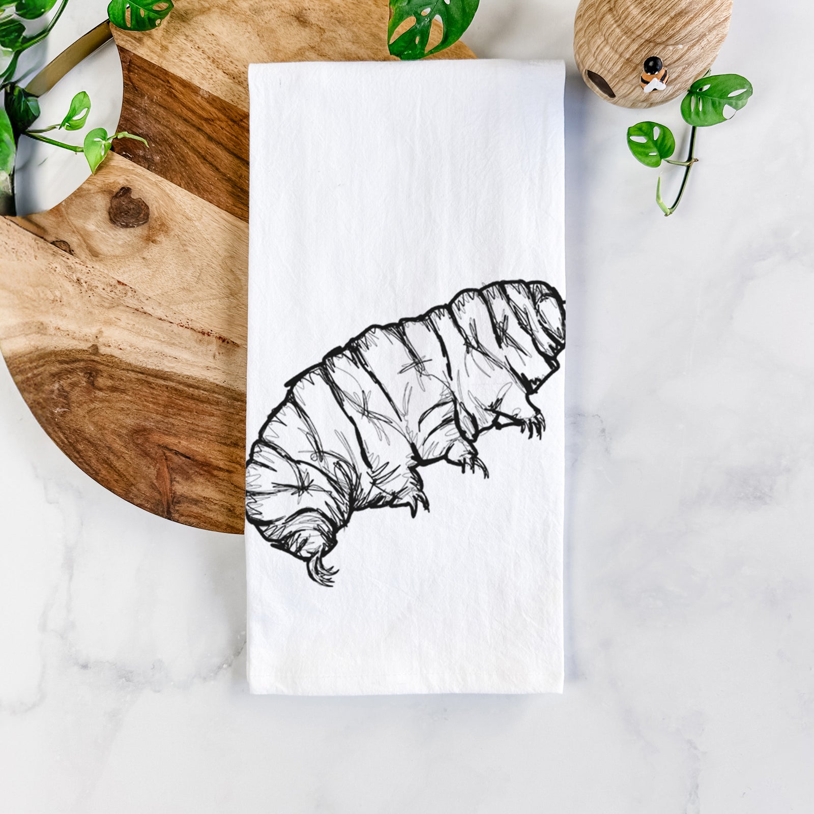 White cotton tea towel printed with a detailed black ink illustration of a tardigrade, or water bear, on a marble counter beside a round wooden cutting board and greenery.