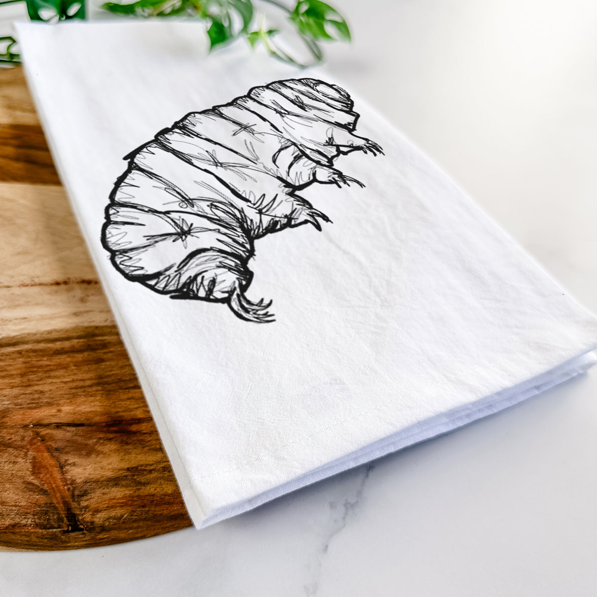 Close-up of a folded white tea towel on a wooden board, showing a detailed black ink illustration of a tardigrade, or water bear.
