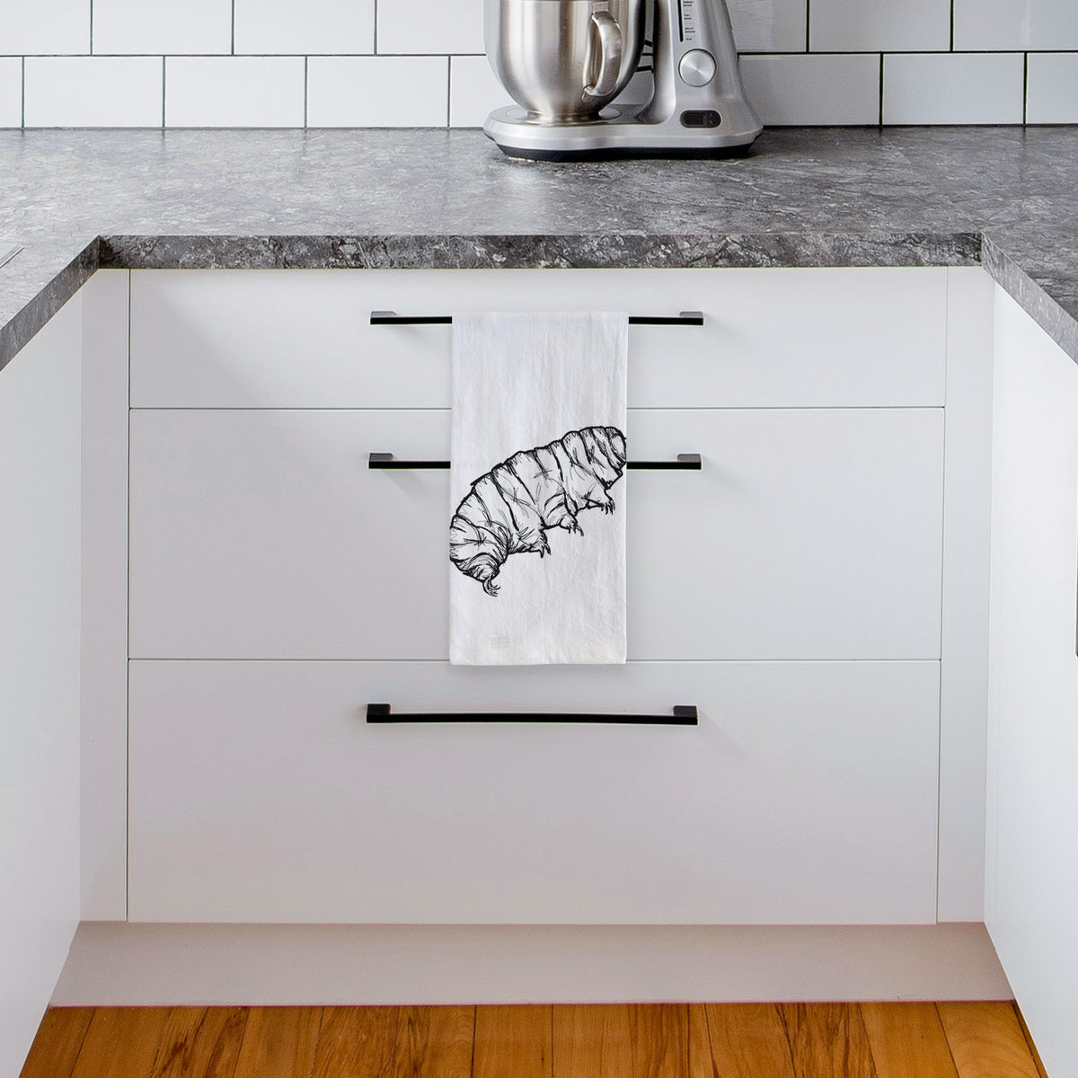 White tea towel printed with a detailed black ink illustration of a tardigrade, or water bear, hanging from a drawer handle in a bright white kitchen.