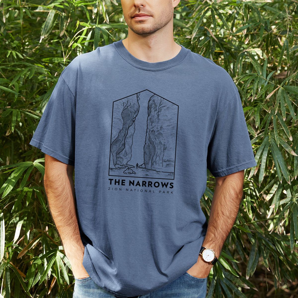 Man wearing a Comfort Colors t-shirt printed with a black line art badge of the tall canyon walls of The Narrows, with the words The Narrows, outdoors.