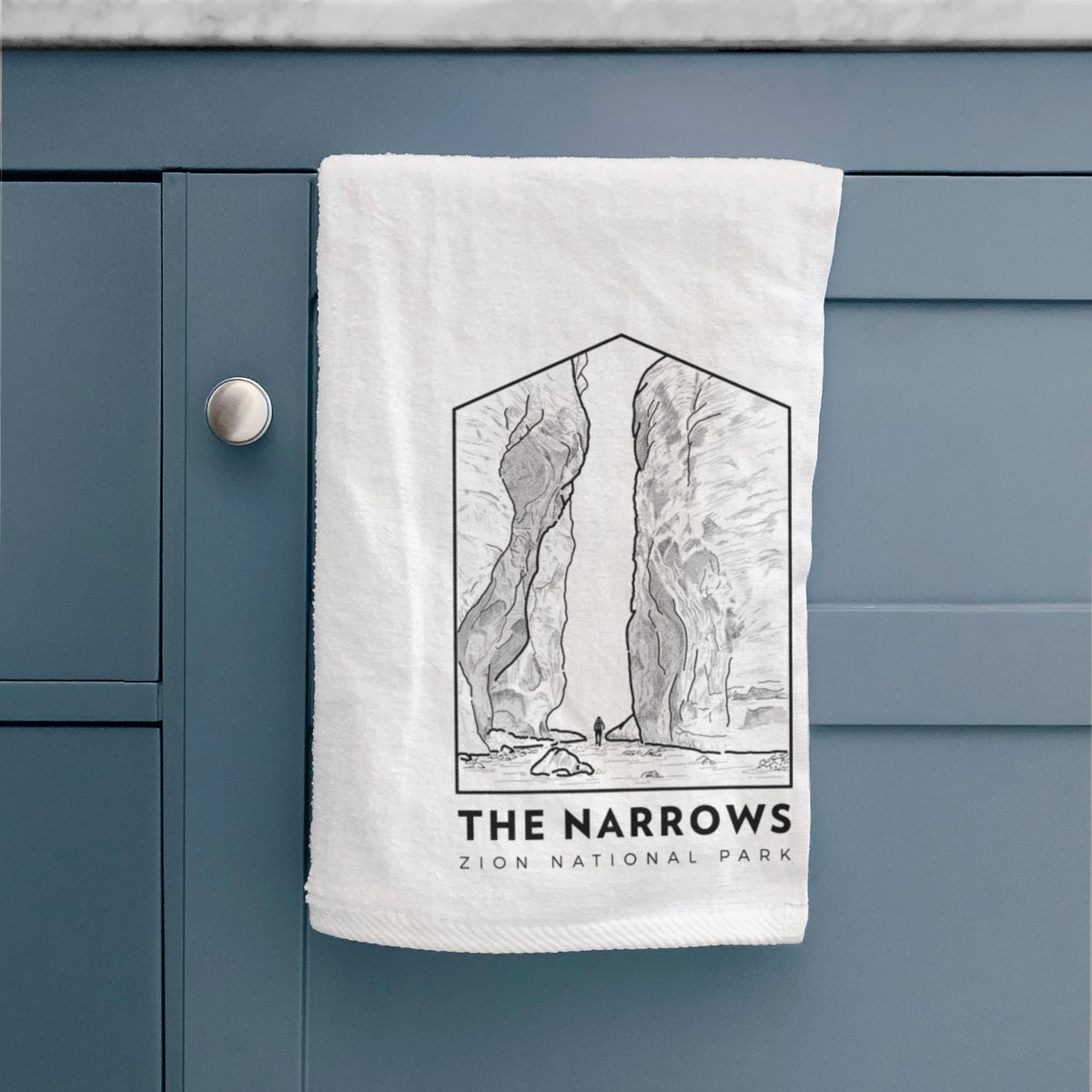 White hand towel printed with a black line art badge of the tall canyon walls of The Narrows, with the words The Narrows, hanging on a cabinet door.