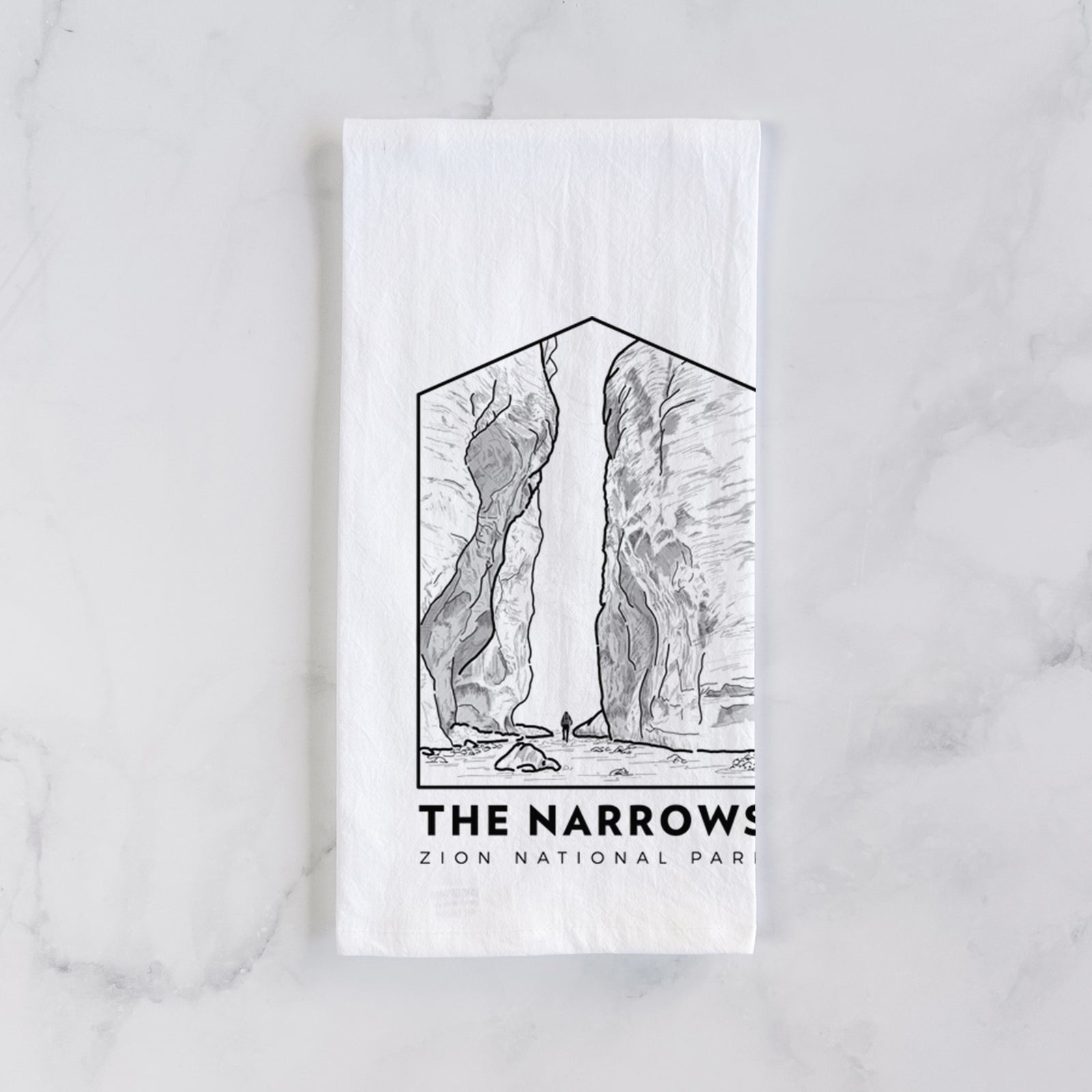 White cotton tea towel printed with a black line art badge of the tall canyon walls of The Narrows, with the words The Narrows, on a marble counter beside a round wooden cutting board and greenery.