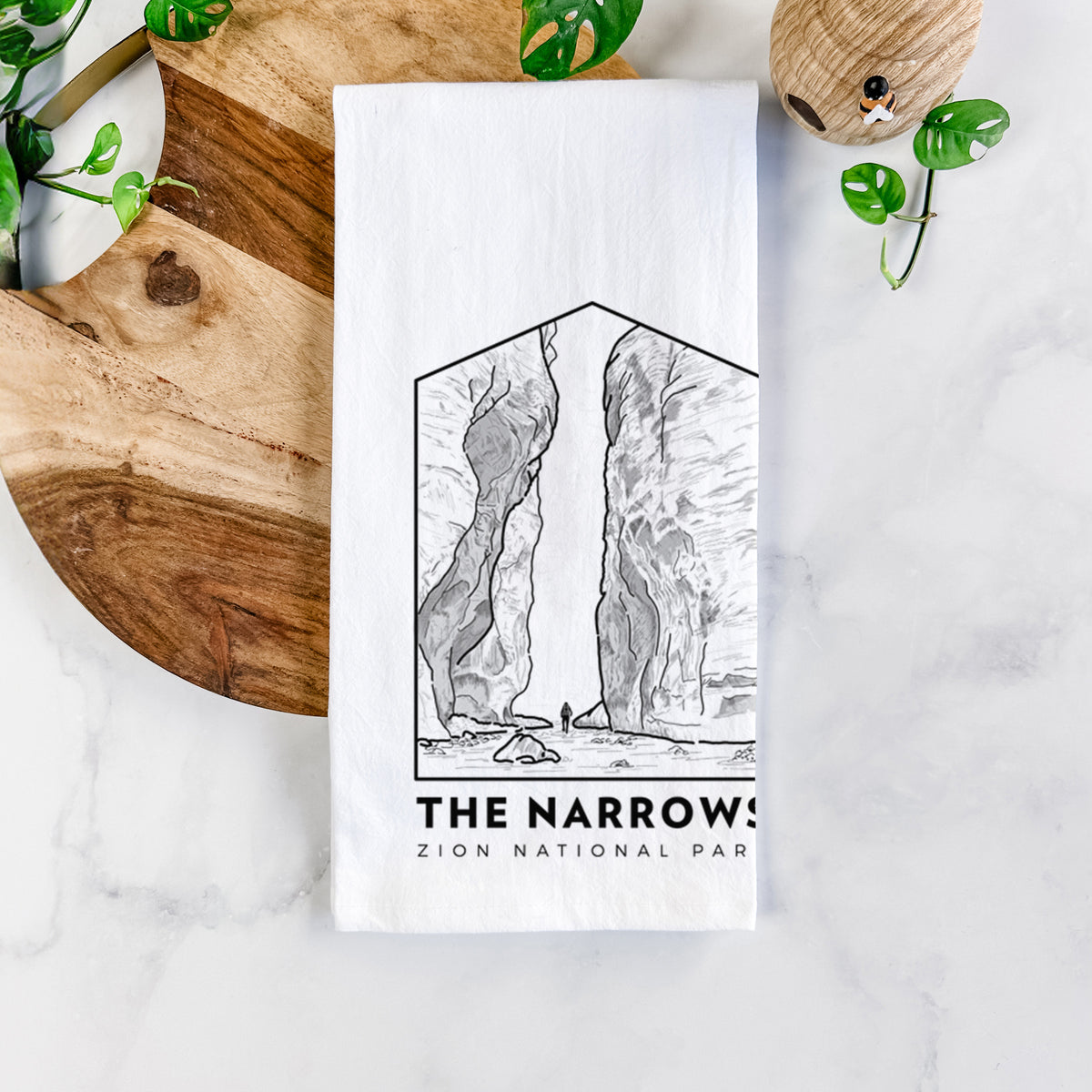 White cotton tea towel printed with a black line art badge of the tall canyon walls of The Narrows, with the words The Narrows, on a marble counter beside a round wooden cutting board and greenery.