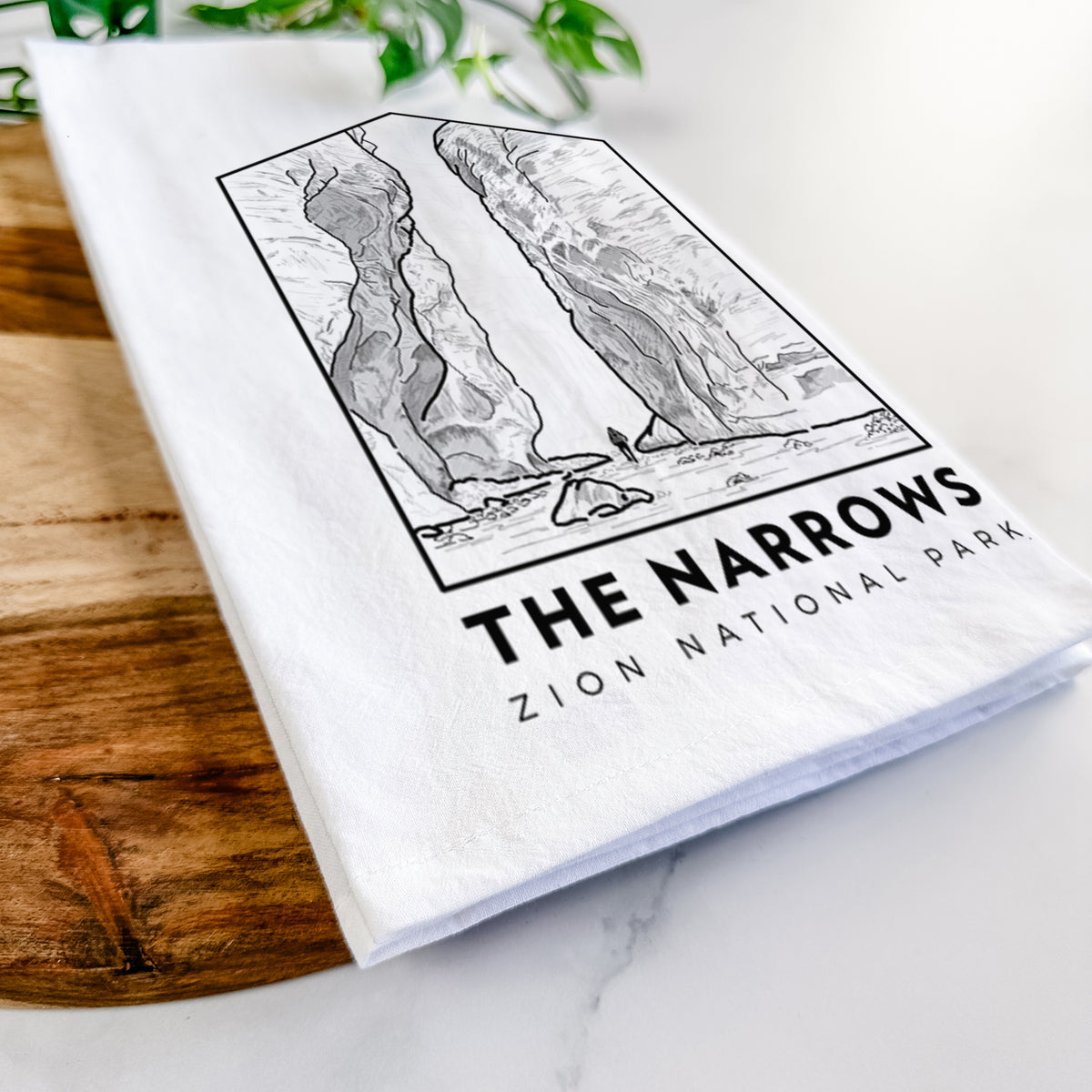 Close-up of a folded white tea towel on a wooden board, showing a black line art badge of the tall canyon walls of The Narrows, with the words The Narrows.